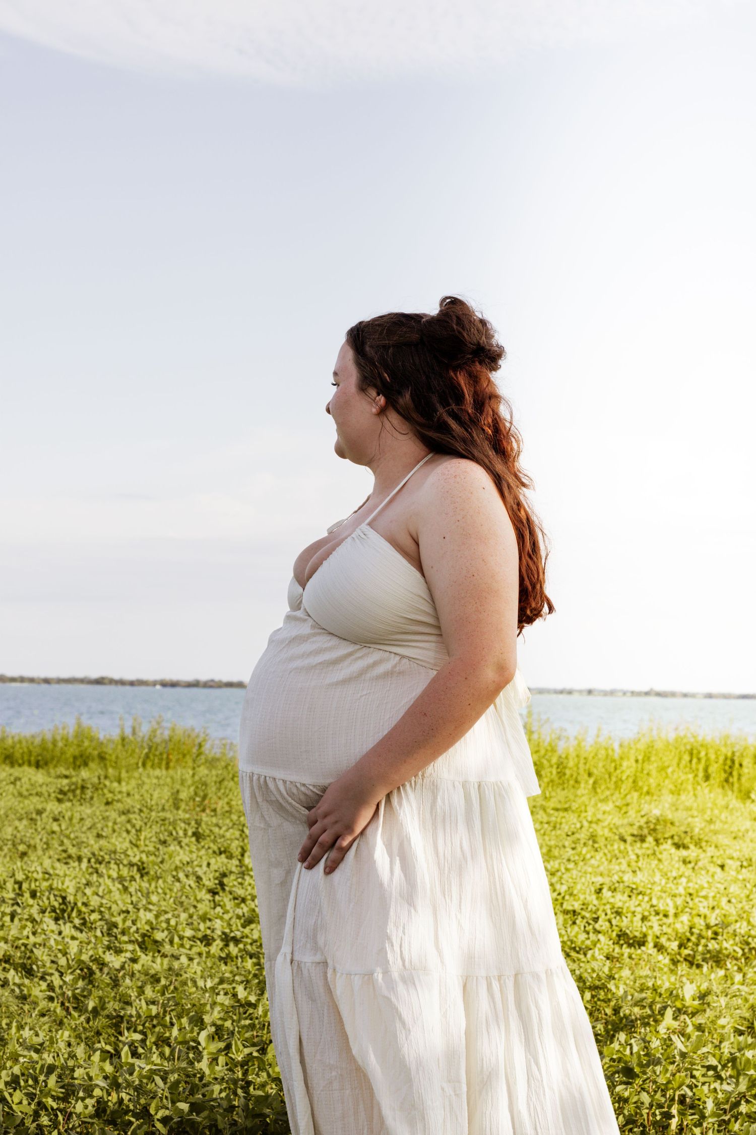 Pregnant woman in white dress standing outdoors near water, cradling her baby bump in a serene green landscape.
