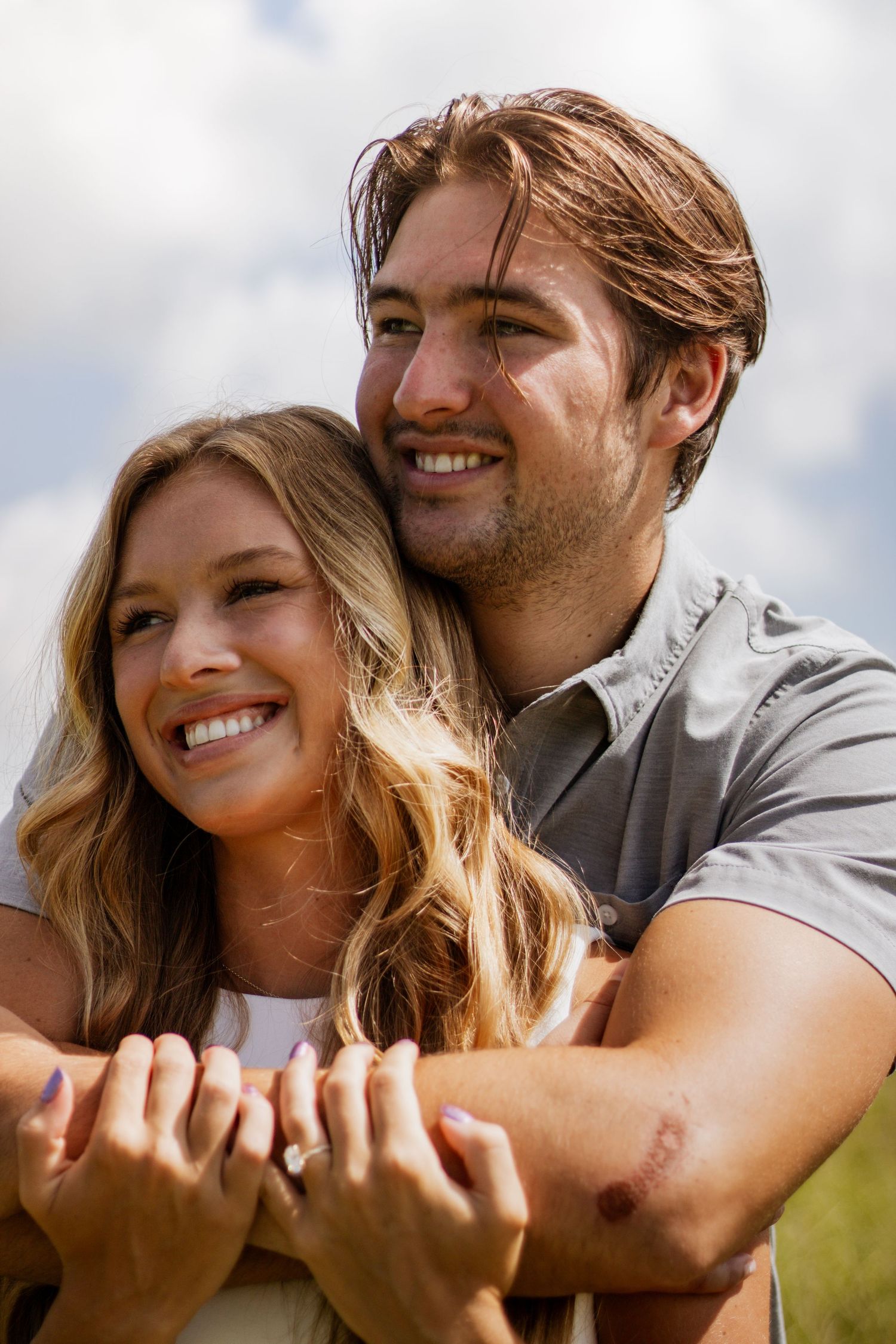 Happy couple embracing outdoors, woman showing engagement ring while man wraps arms around her under cloudy sky.