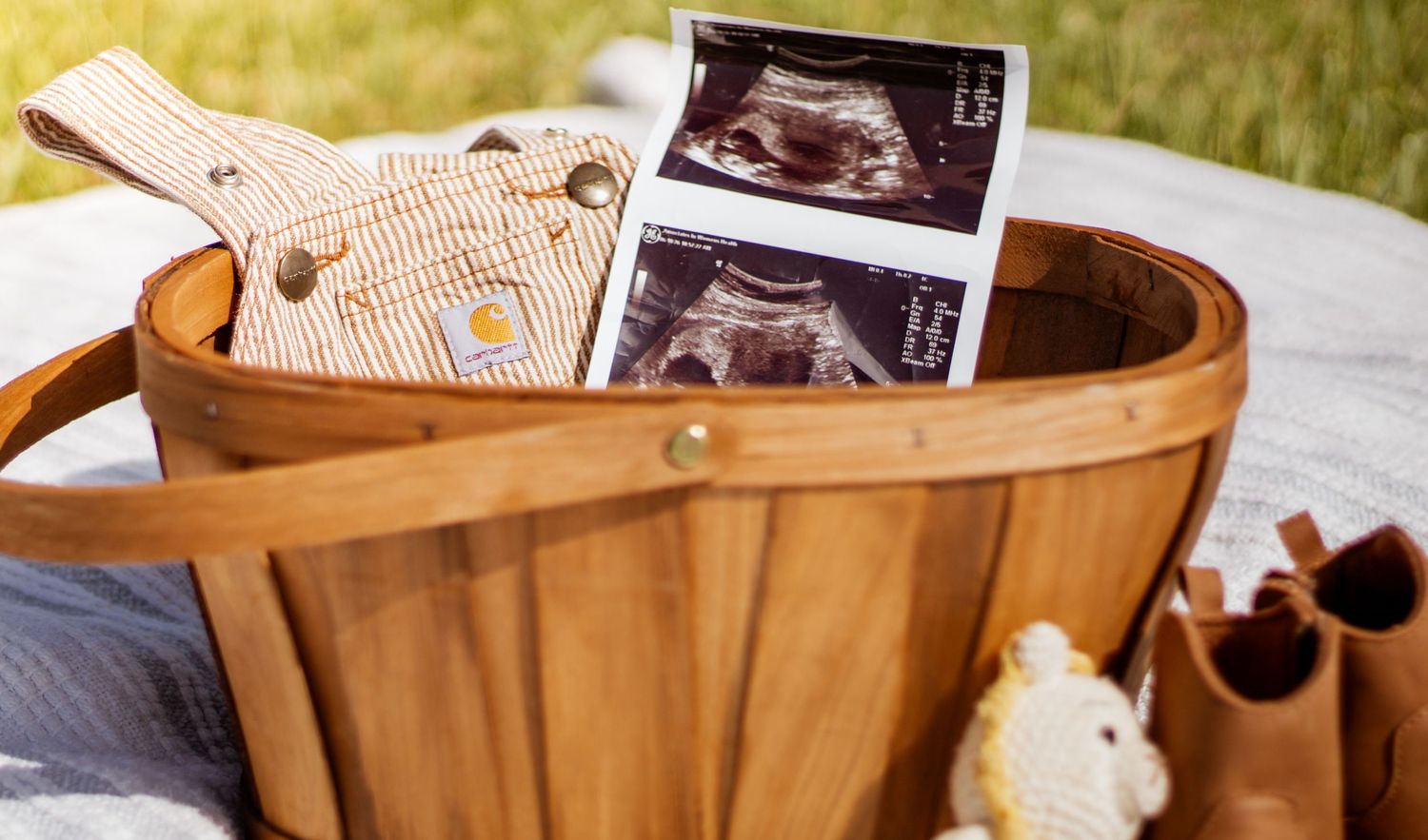 Wooden basket with ultrasound photos, baby bib, and tiny shoes on a blanket outdoors.