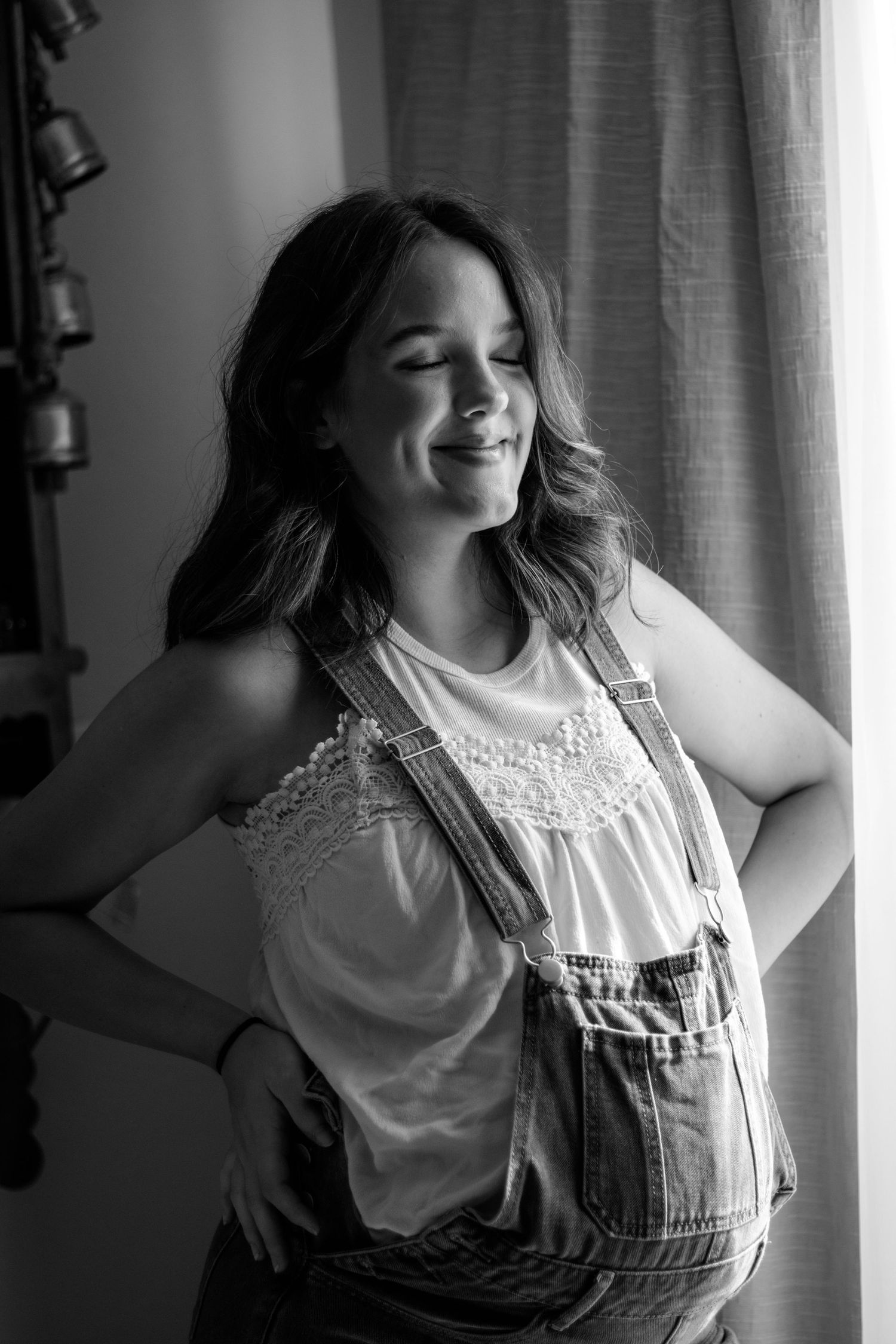 Black and white portrait of a smiling pregnant woman in denim overalls and white lace top standing by a window.