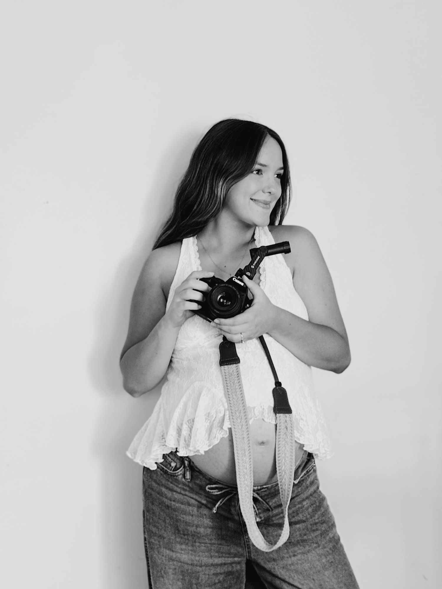 Black and white photo of a young woman holding a DSLR camera, wearing a white top and jeans, smiling softly.