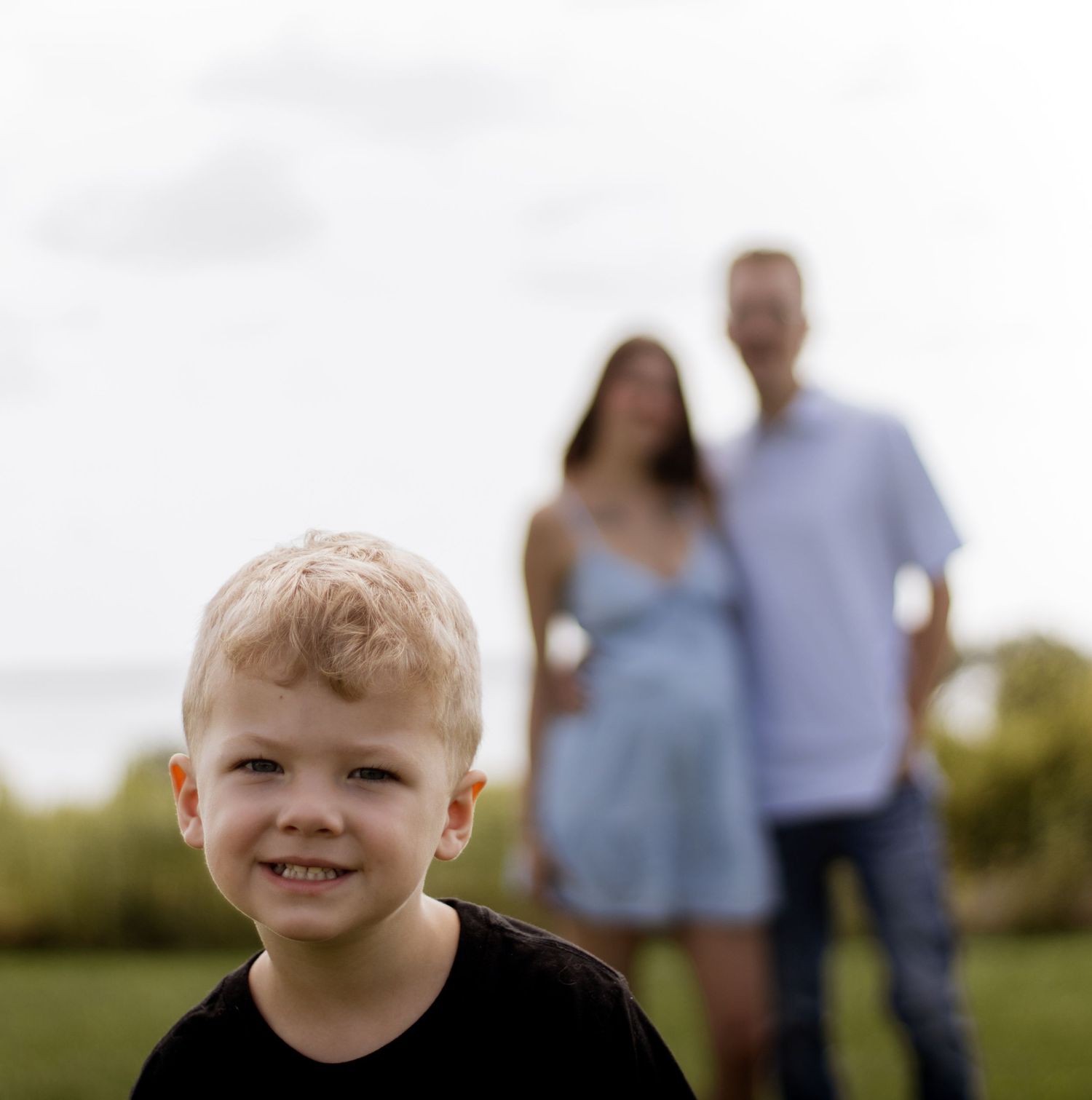 A smiling blond boy in a black shirt poses in focus while his parents stand blurred in a green field behind him.