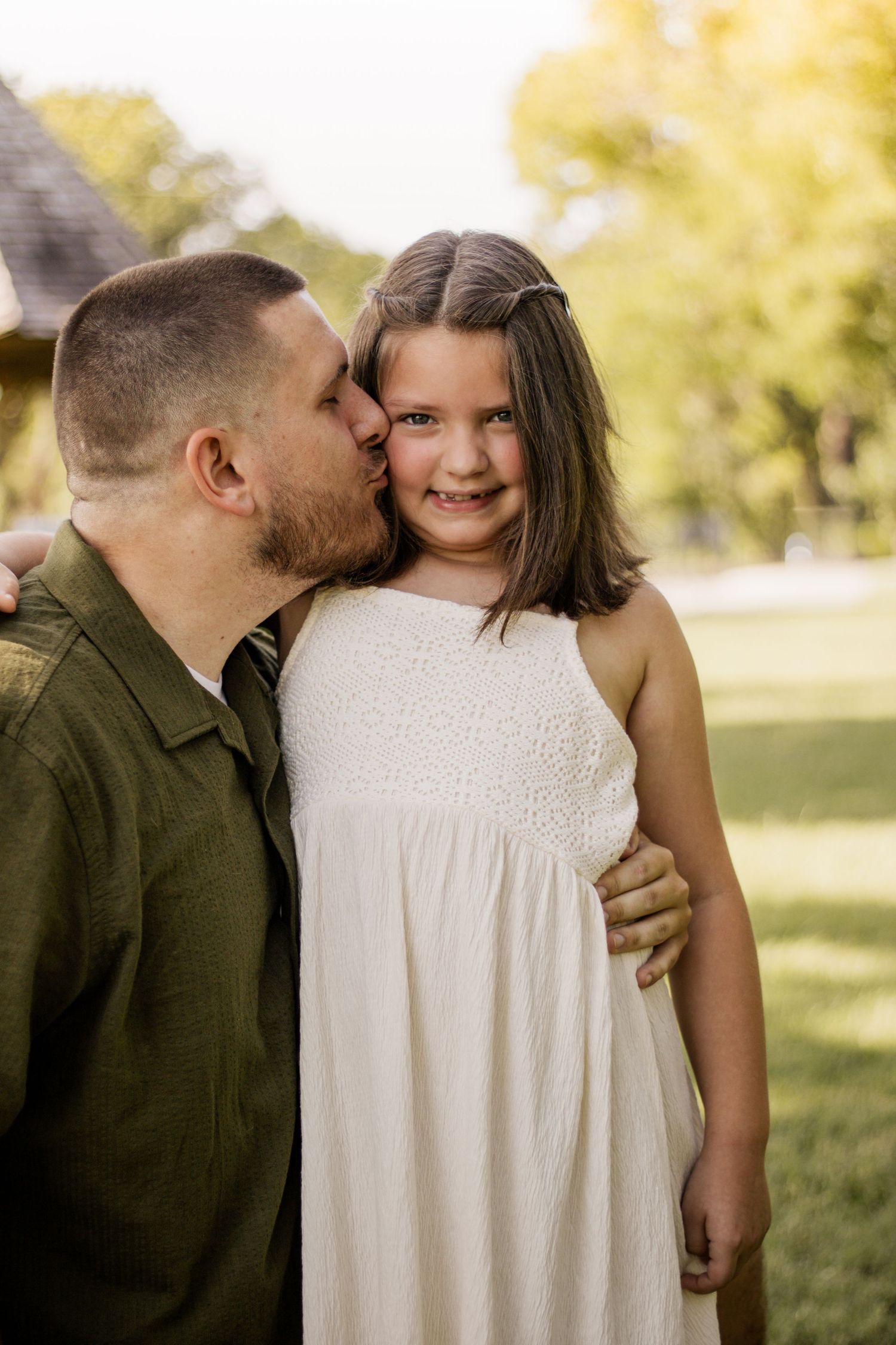 A father kisses his smiling young daughter on the cheek outdoors, she wears a white dress in a green park setting.