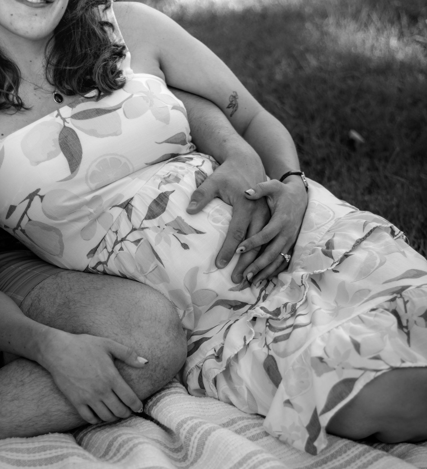 Black and white photo of a pregnant couple sitting outdoors, both cradling the baby bump tenderly.