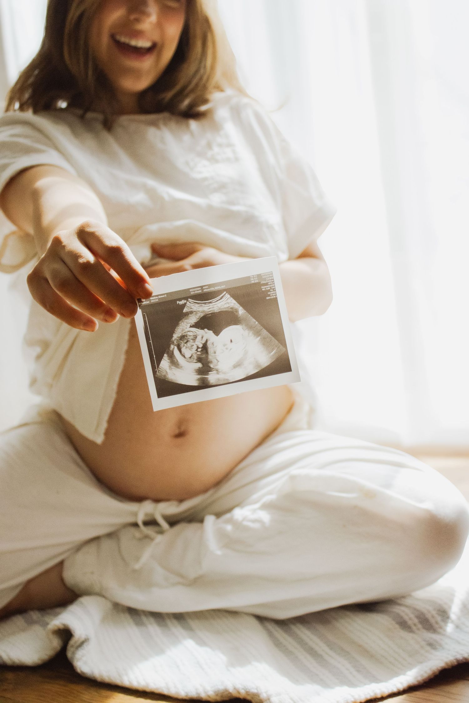 Happy pregnant woman laughing while holding ultrasound photo near bare baby bump indoors.