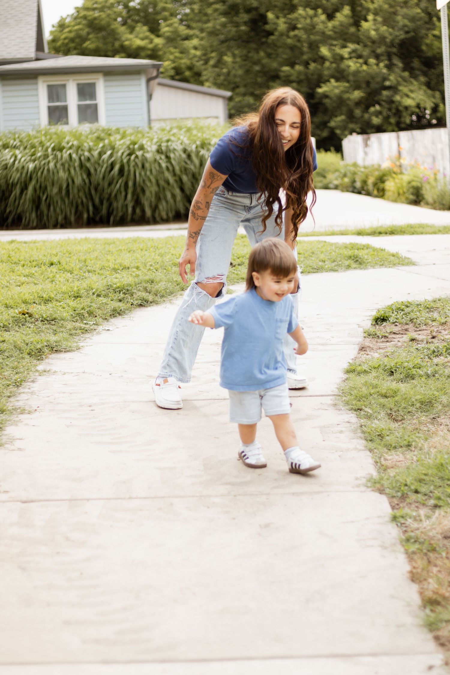 Smiling mother encouraging toddler to walk on concrete path outdoors.