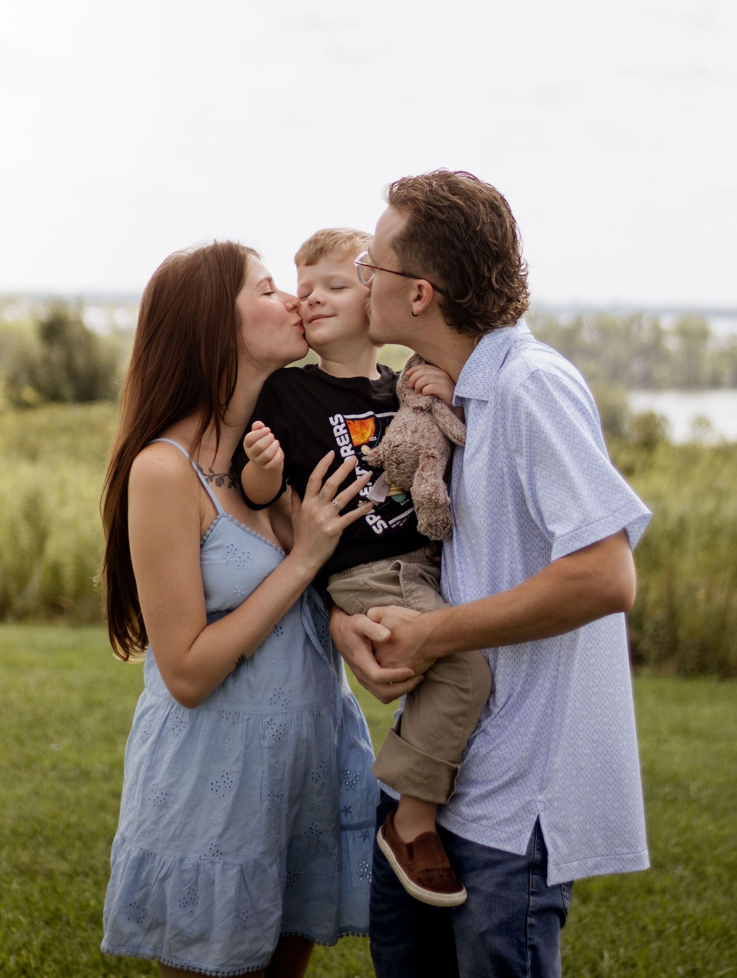 A young family shares kisses with their toddler boy outdoors near a lake on a green lawn.