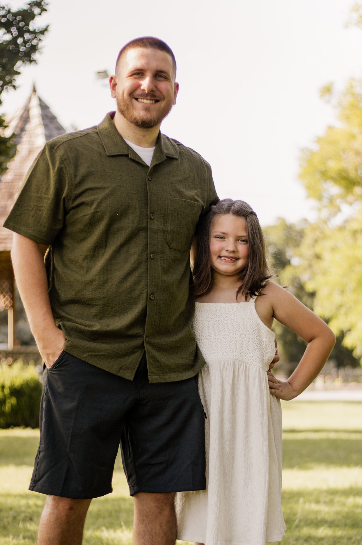 A smiling man in a green shirt and a young girl in a white dress pose together outdoors on a sunny day.