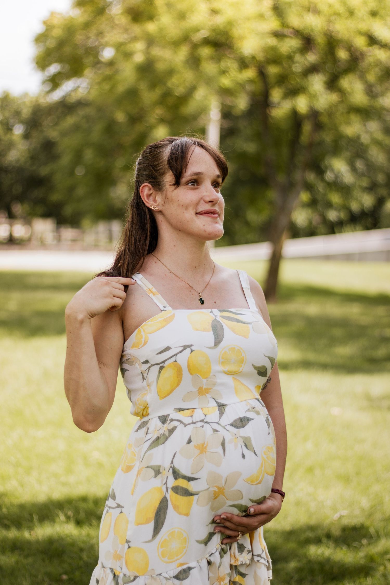 Pregnant woman in floral lemon print dress cradling baby bump outdoors in green park setting.