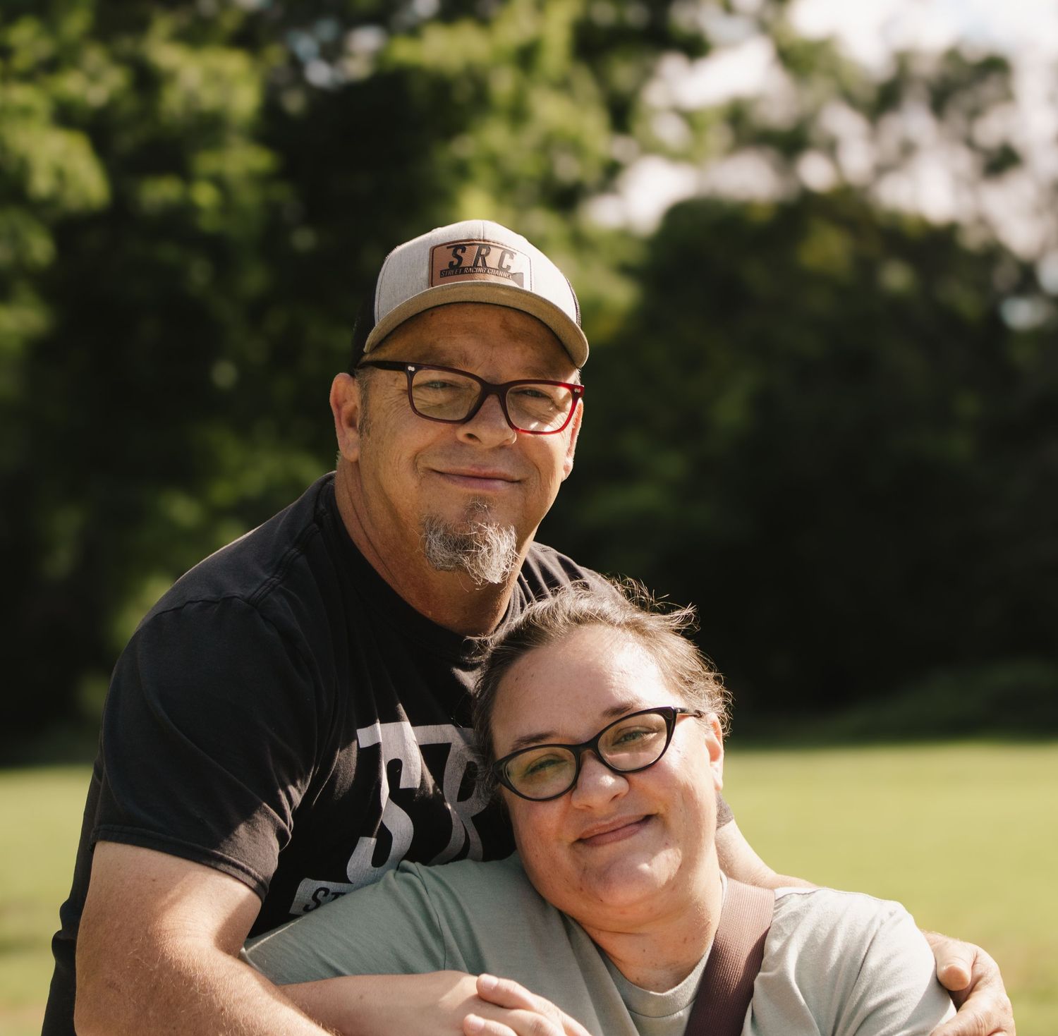 Couple posing closely together outdoors, smiling at camera with green trees in the background.