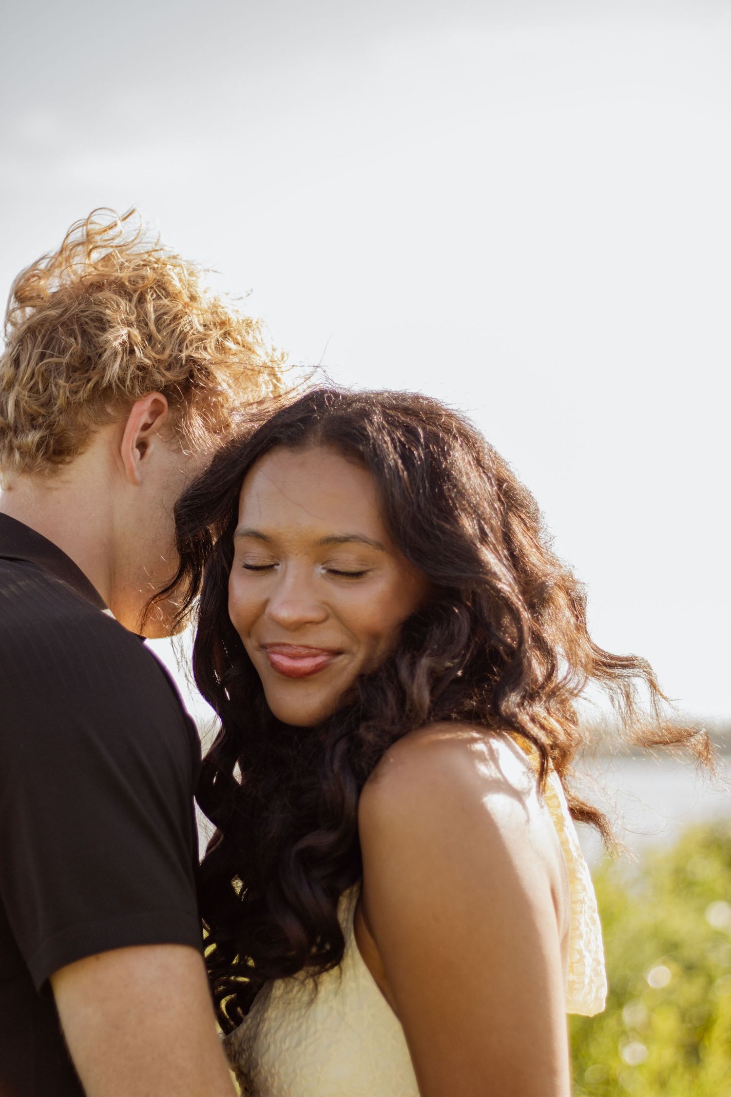 A curly-haired man whispers to a smiling mixed-race woman outdoors in warm sunlight.