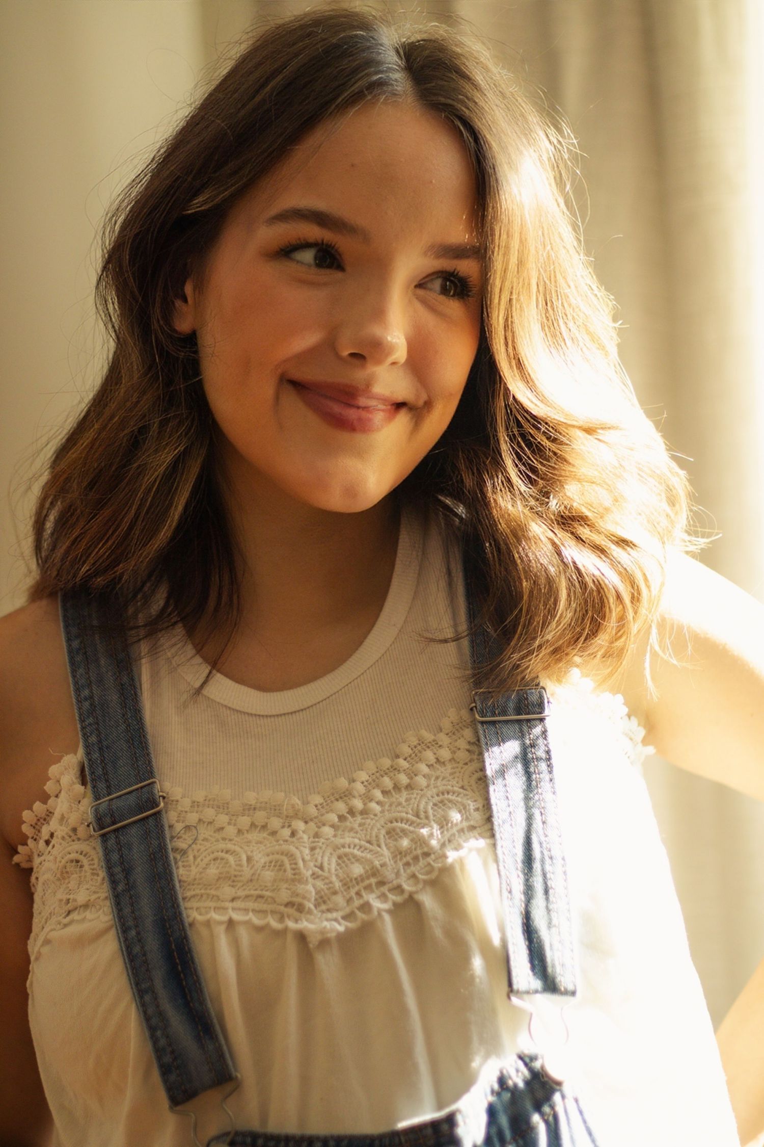 Young girl with wavy brown hair smiling, wearing denim overalls over a white lace top in warm sunlight.