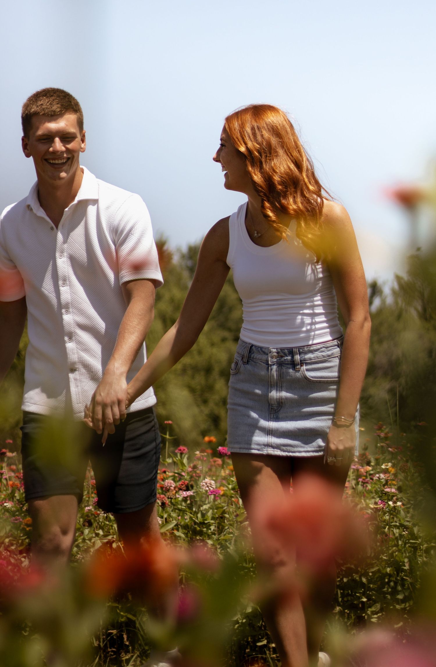 A smiling couple holds hands while walking through a vibrant flower field on a sunny day.
