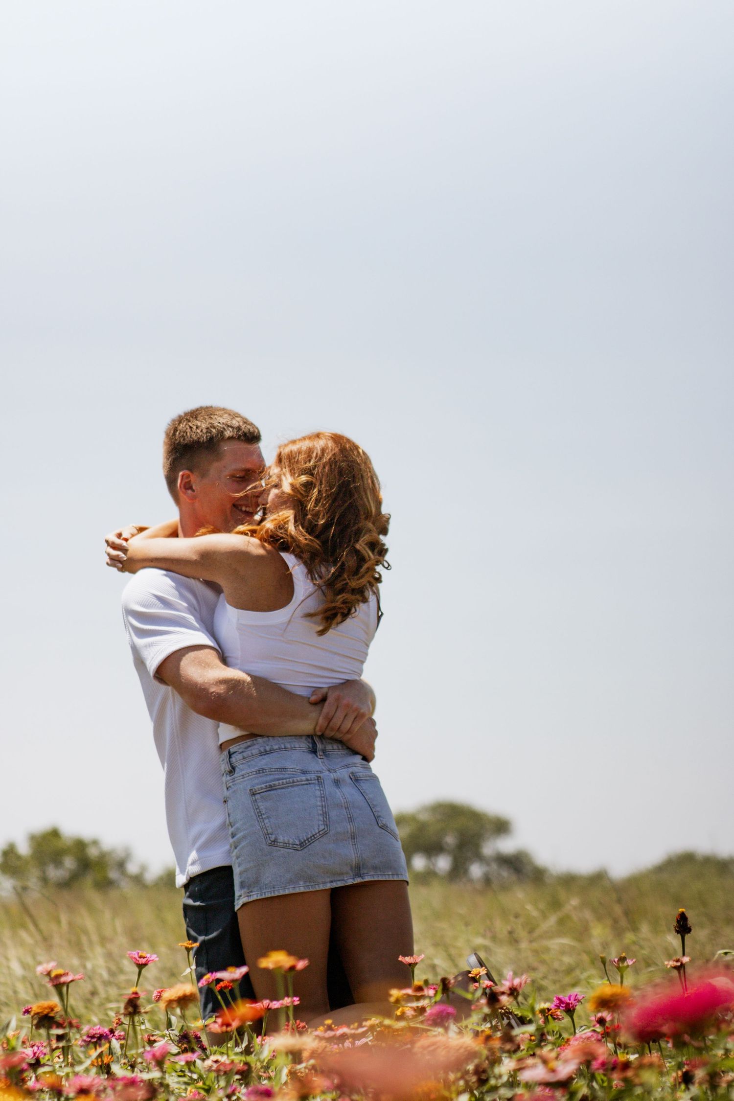 Couple embracing and kissing in a vibrant flower field under a clear sky on a sunny day.