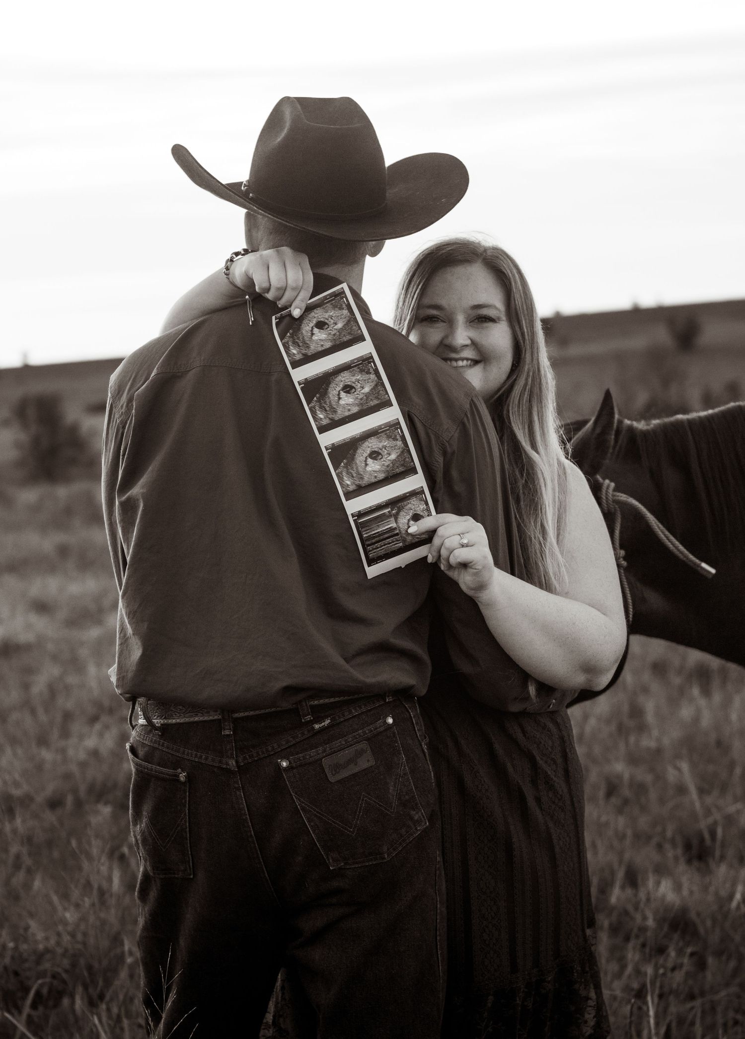 Couple holding ultrasound photos in a field, man in cowboy hat and jeans, woman smiling beside a horse, black and white photo.