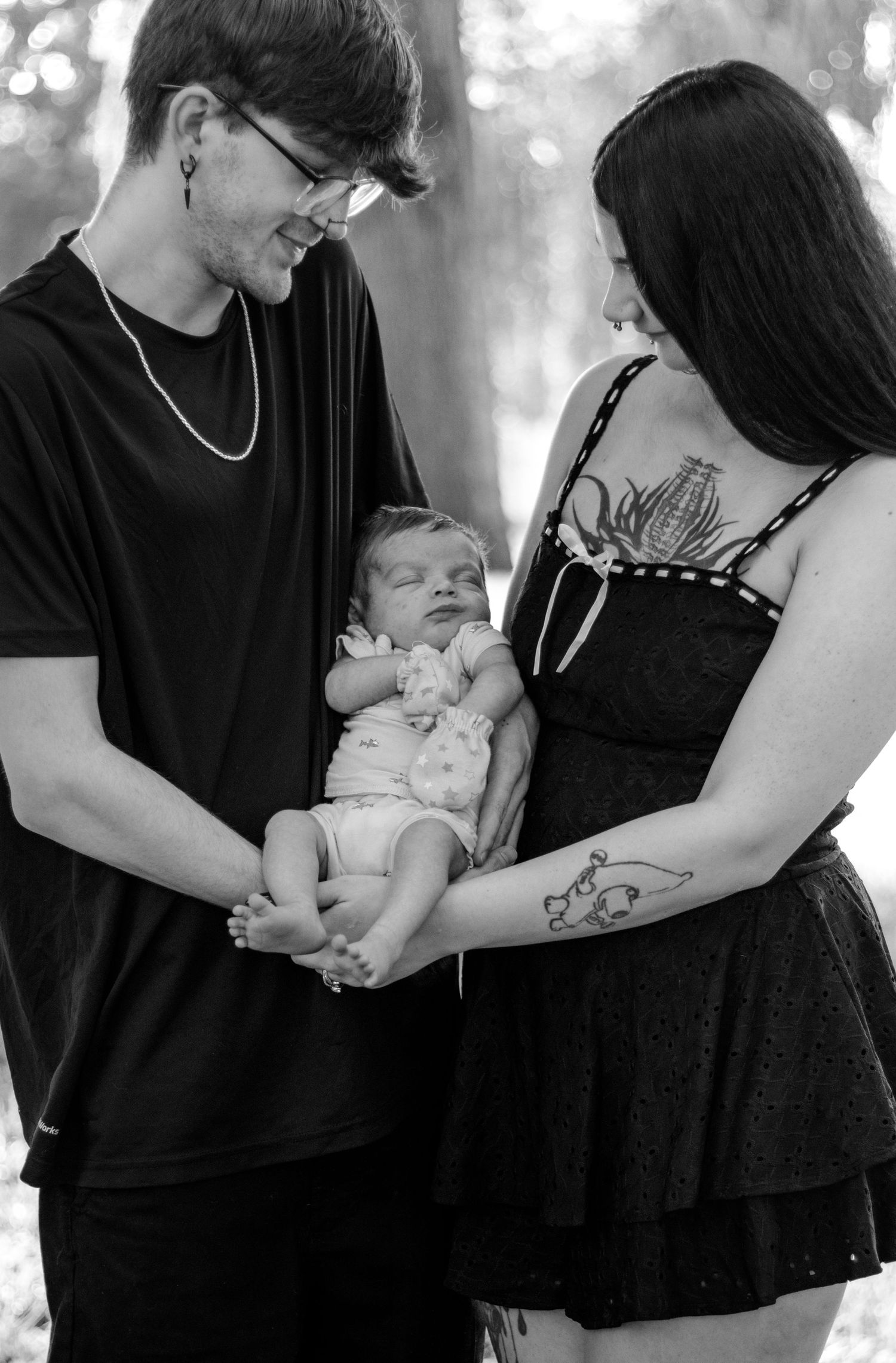 Black and white photo of young tattooed parents lovingly holding their newborn baby outdoors.