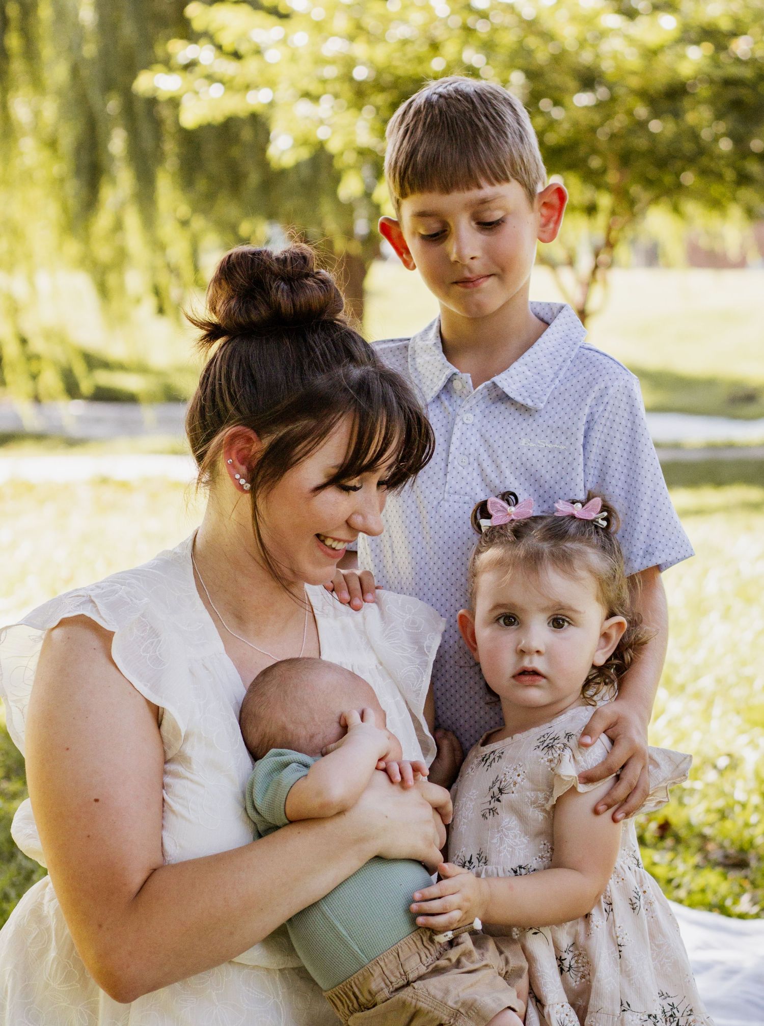 A mother holds her newborn while her two young children gather close in a sunny outdoor family portrait.