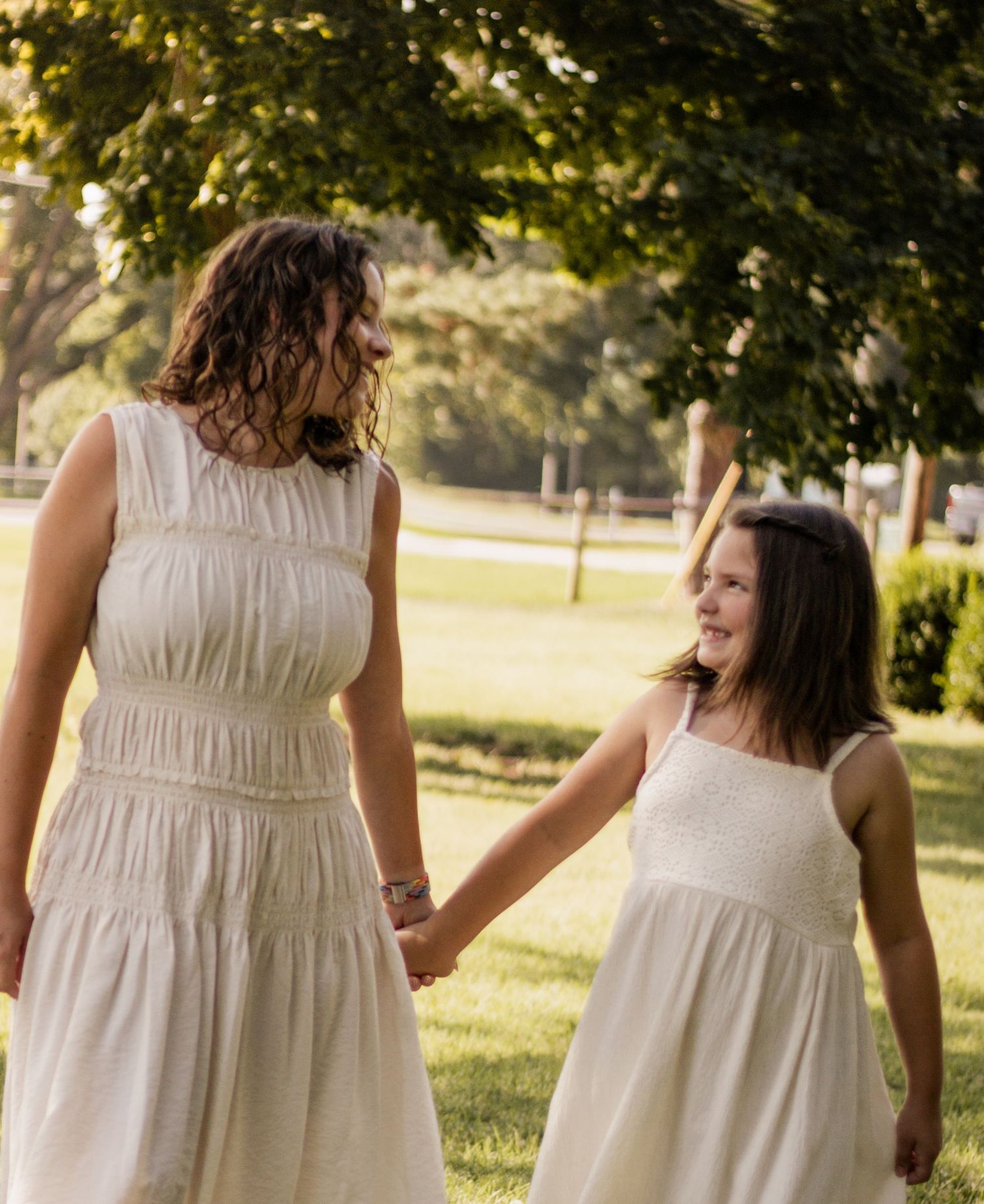 A mother and daughter in matching white dresses hold hands and smile at each other in a sunny park.