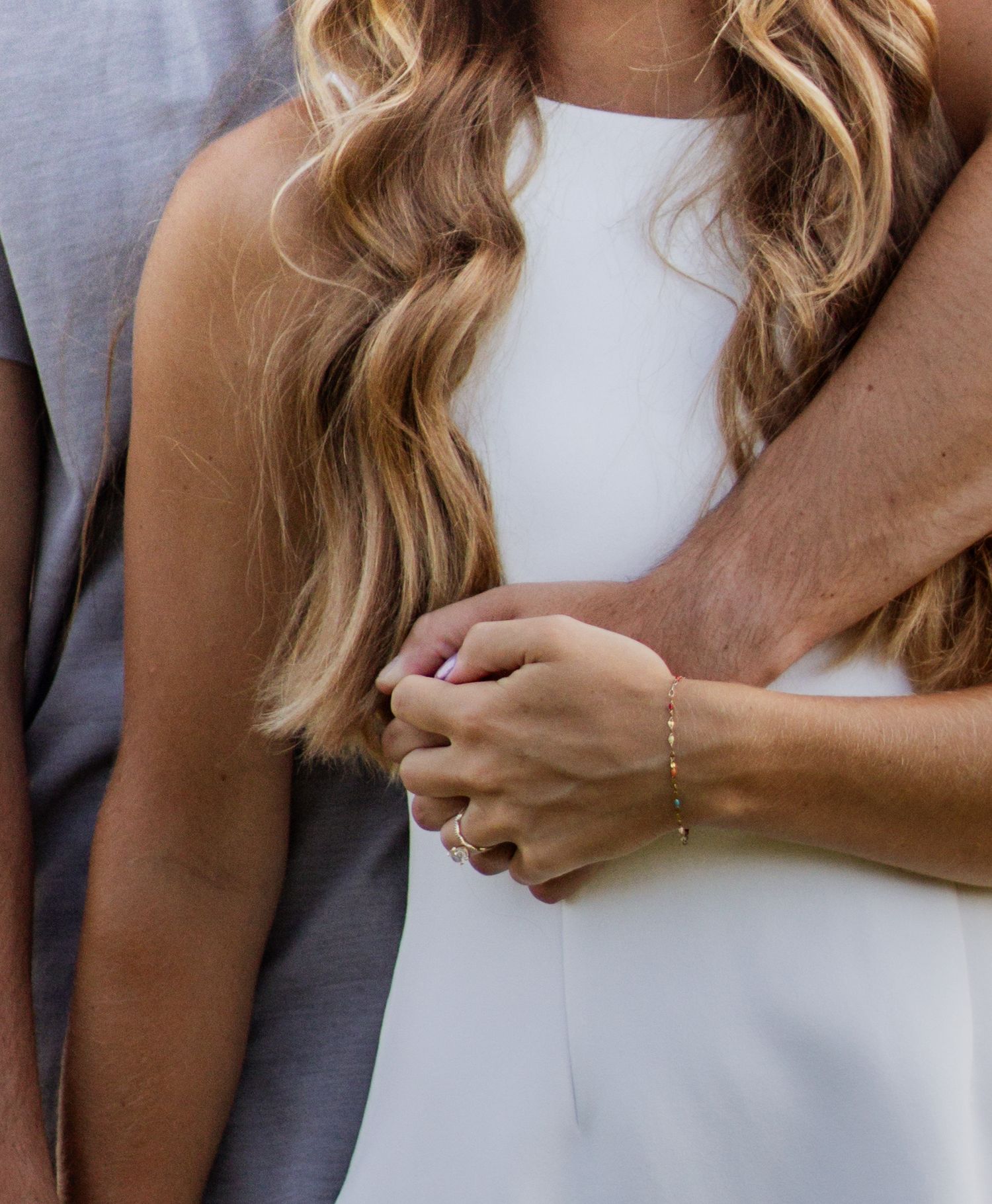 Couple embracing in white dress and gray shirt, hands clasped together with delicate gold bracelet and engagement ring.