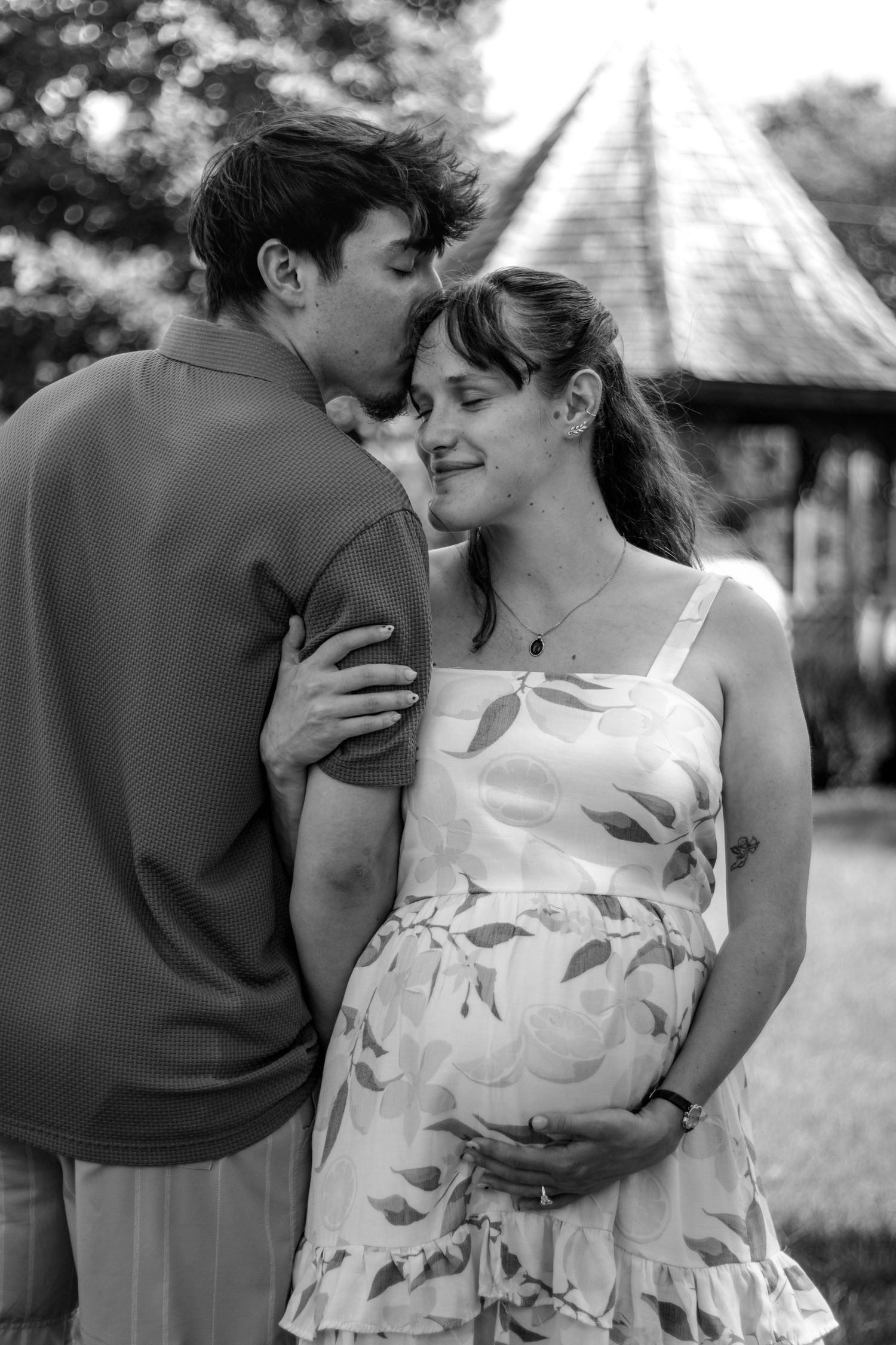 Black and white photo of a couple sharing a tender moment, with the woman cradling her pregnant belly outdoors.