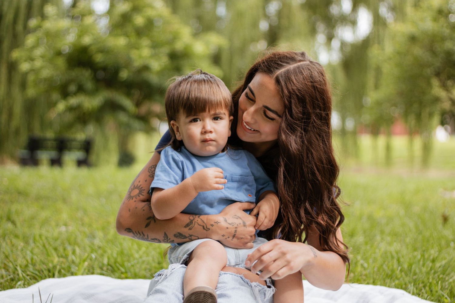 A young mother with tattoos sits on a blanket in a park, lovingly holding her toddler son in a blue shirt.