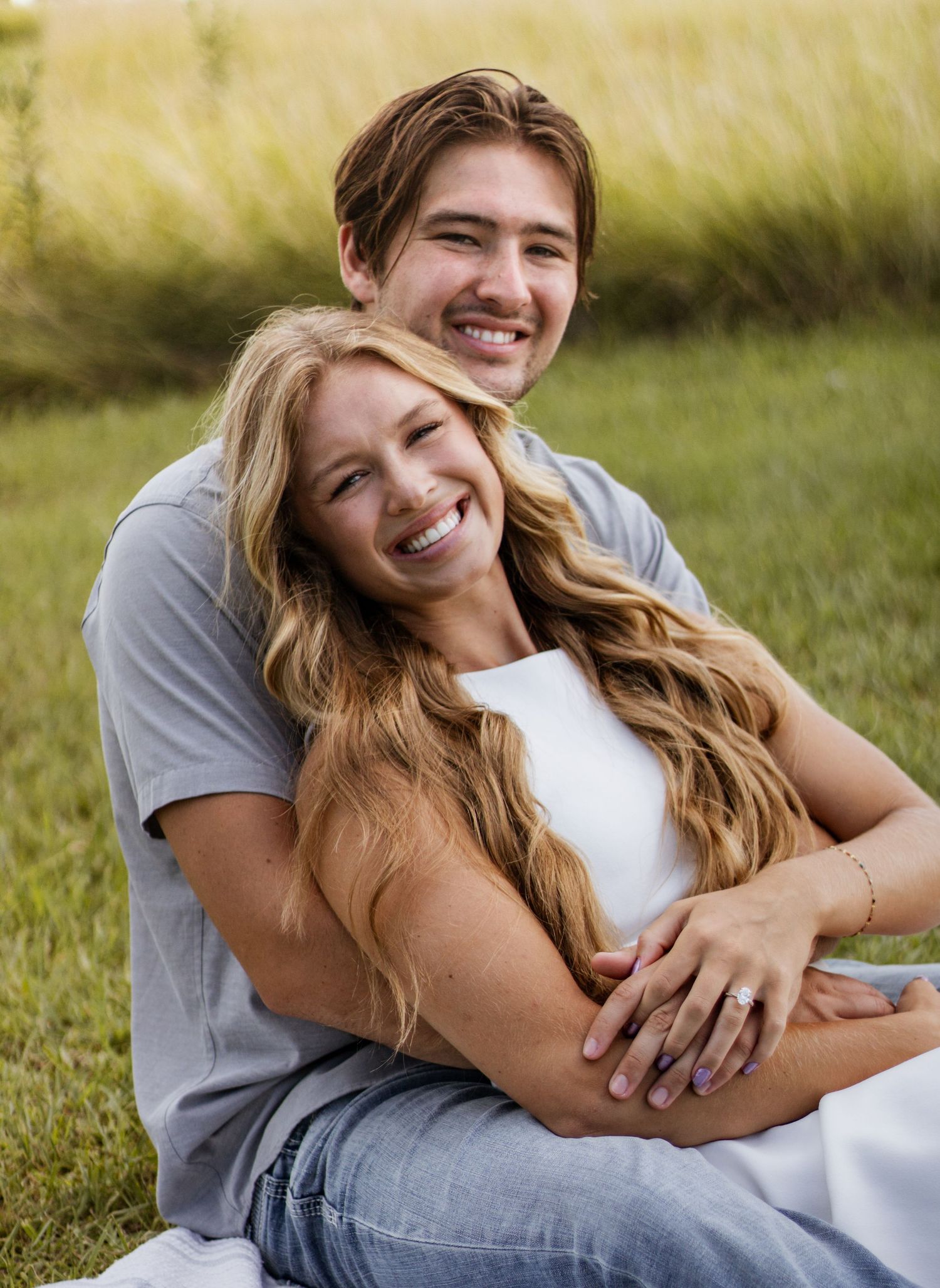 A smiling couple sits together in a green field, the woman with long blonde hair leaning back into the man's embrace.