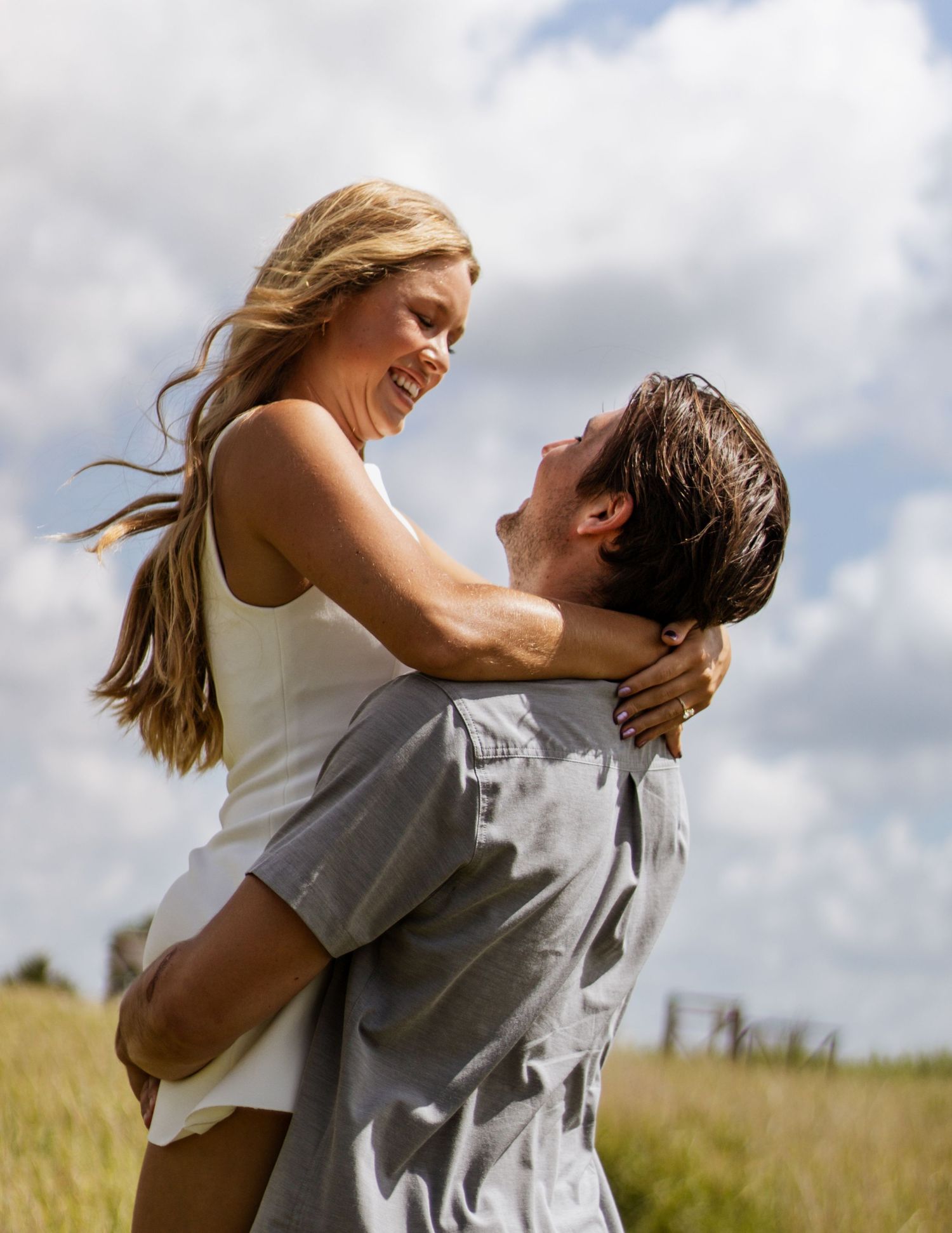 A joyful couple embraces in a sunny field, man lifting smiling woman in white dress with cloudy sky backdrop.