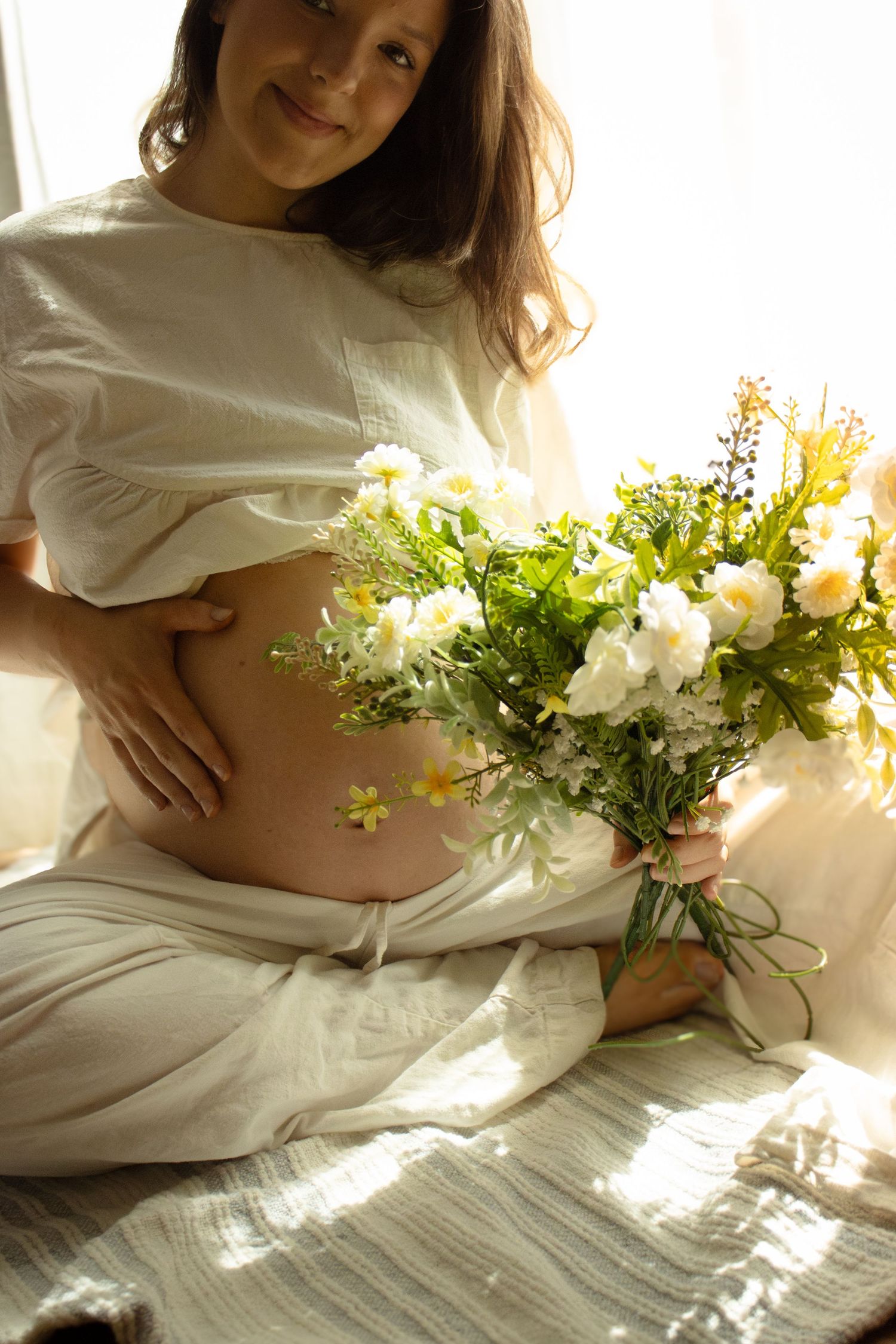 Smiling pregnant woman in white outfit holding bouquet of white and yellow wildflowers while cradling her baby bump.