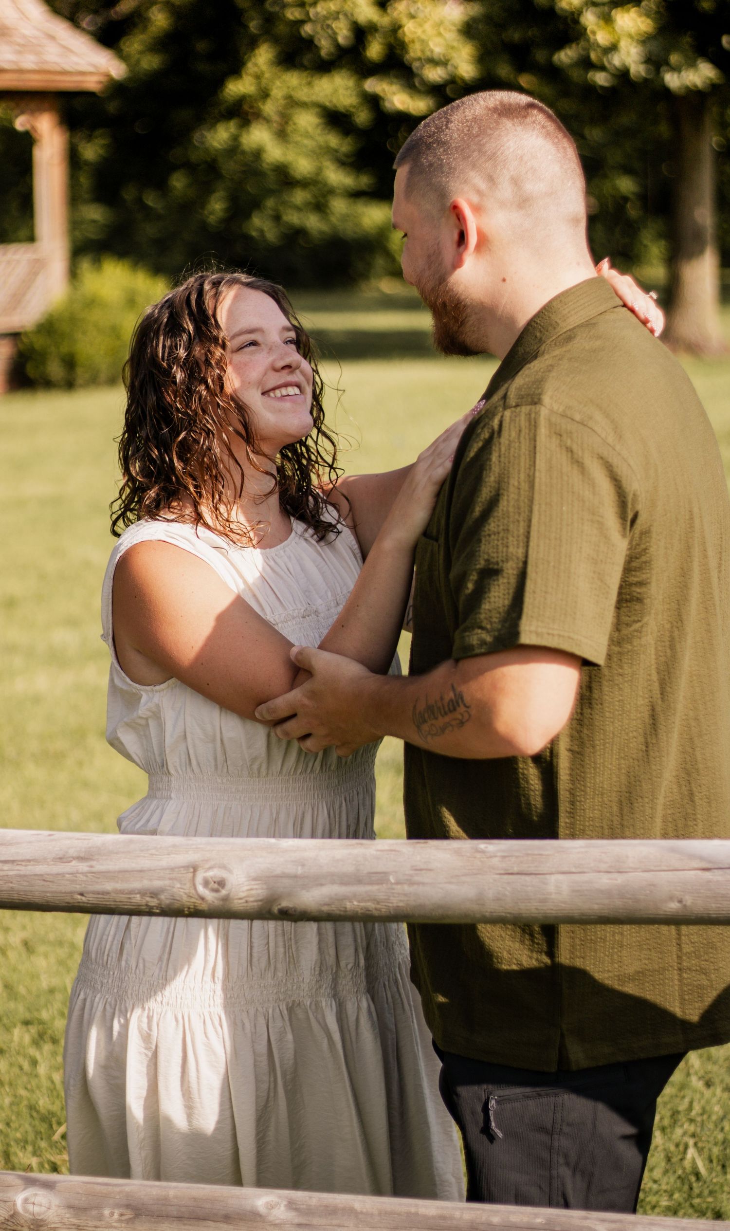 A smiling couple in a sunny park, woman in white dress and man in olive shirt, standing by a wooden fence.