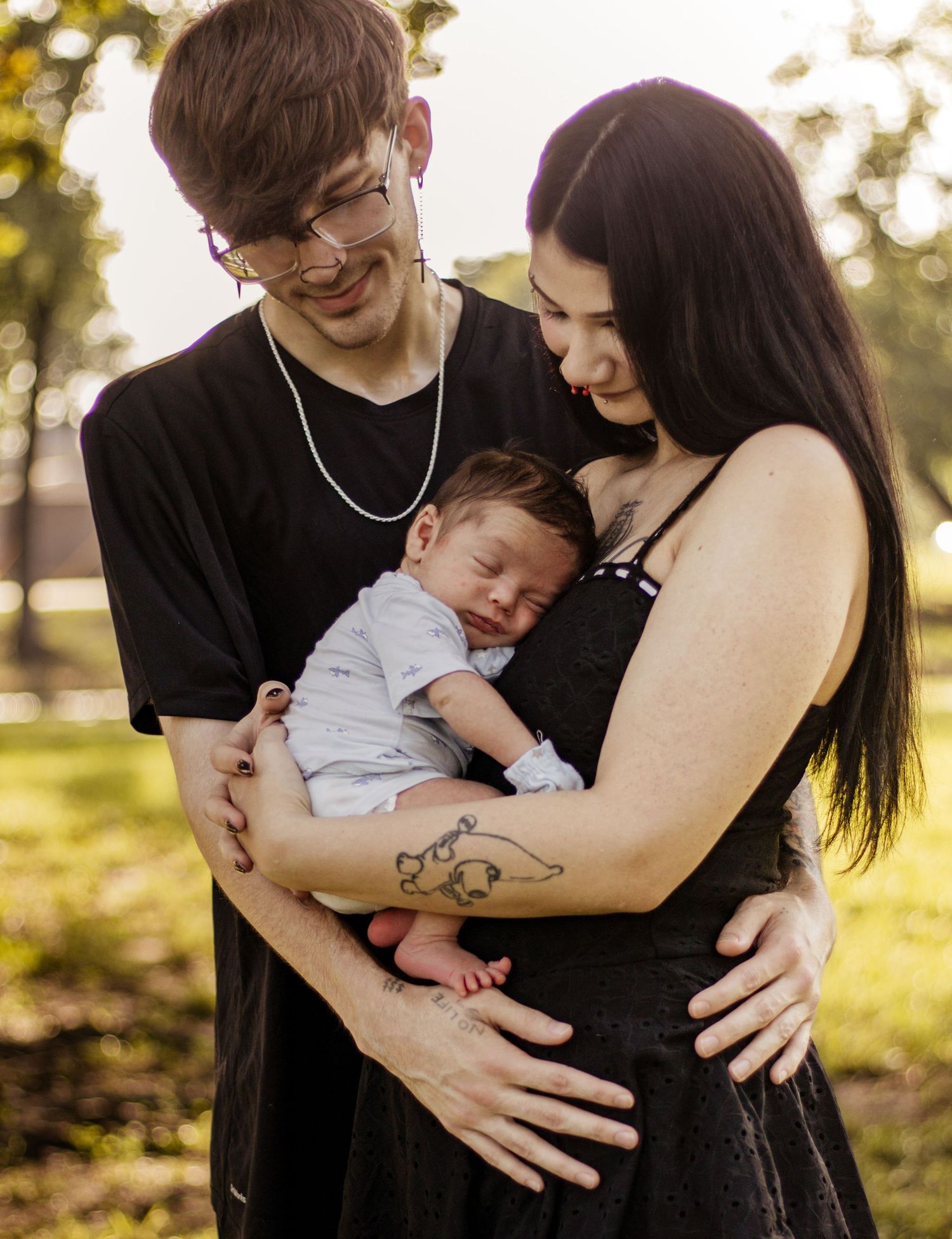 Young couple in black outfits holding their sleeping newborn baby outdoors in golden hour sunlight.