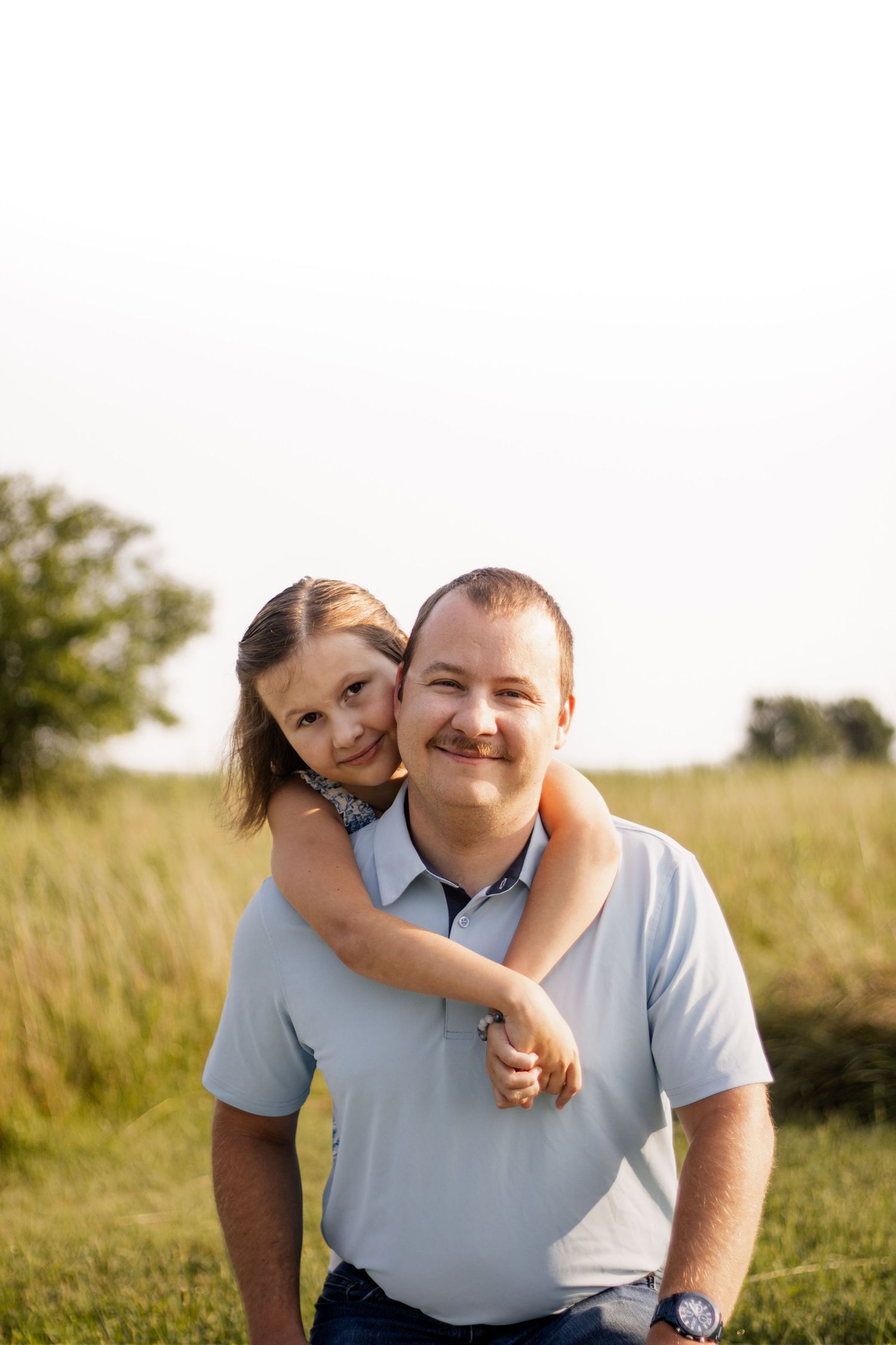 A smiling man in a light blue polo shirt gives a young girl a piggyback ride in a sunny green field.
