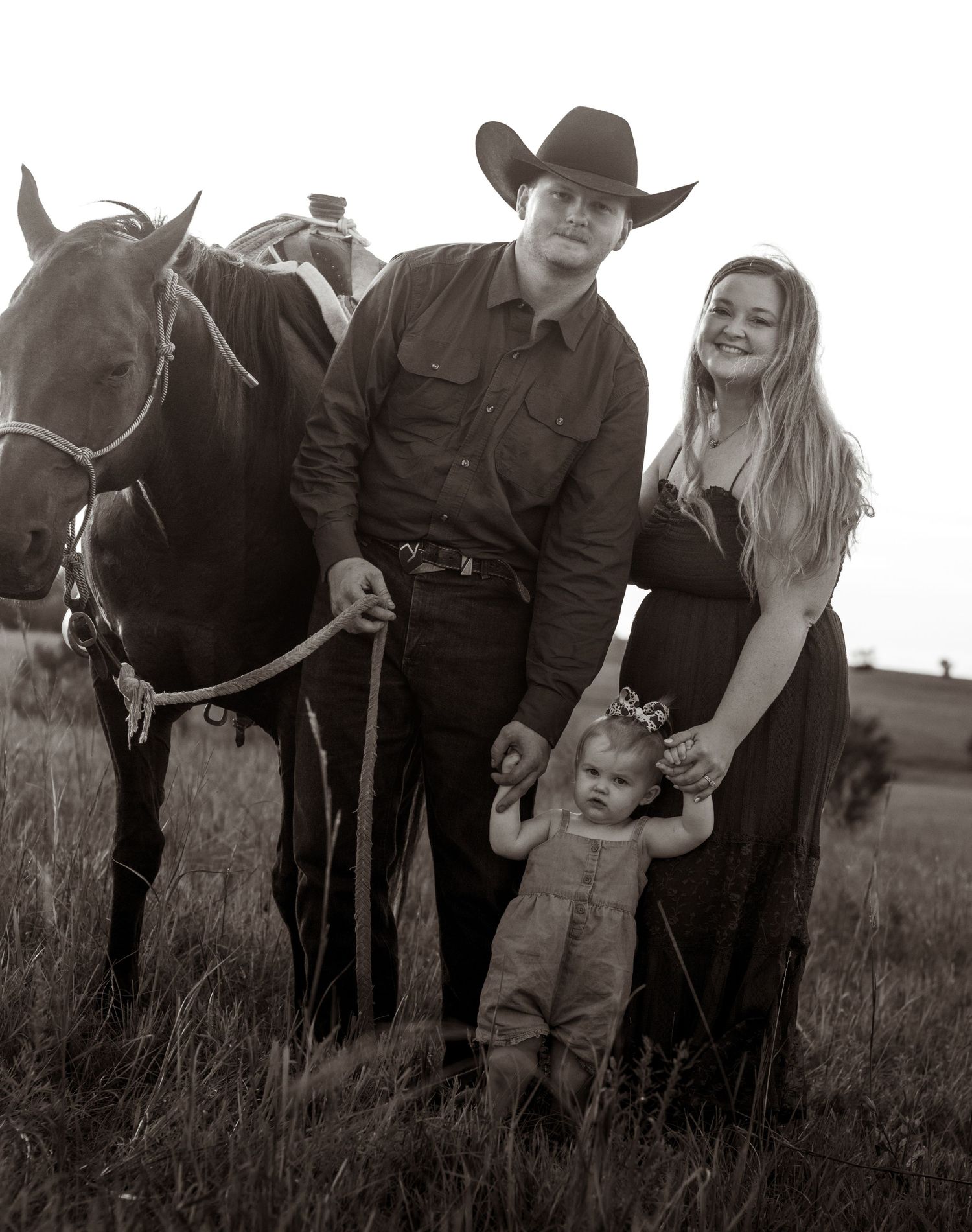 Black and white photo of a cowboy, woman, and toddler posing with a horse in a grassy field.