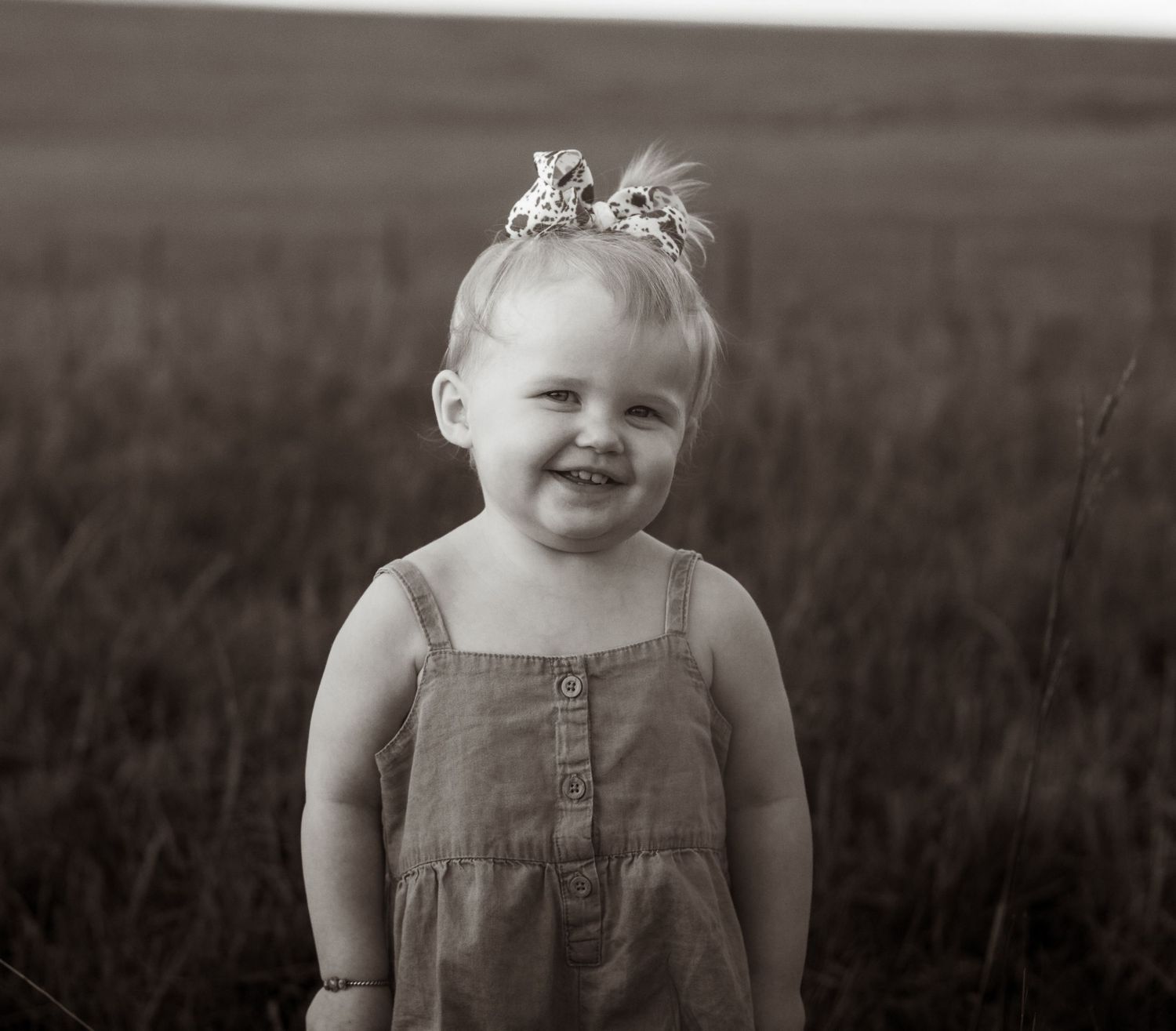Smiling toddler girl in denim dress with bow in hair standing in field, black and white portrait photo.