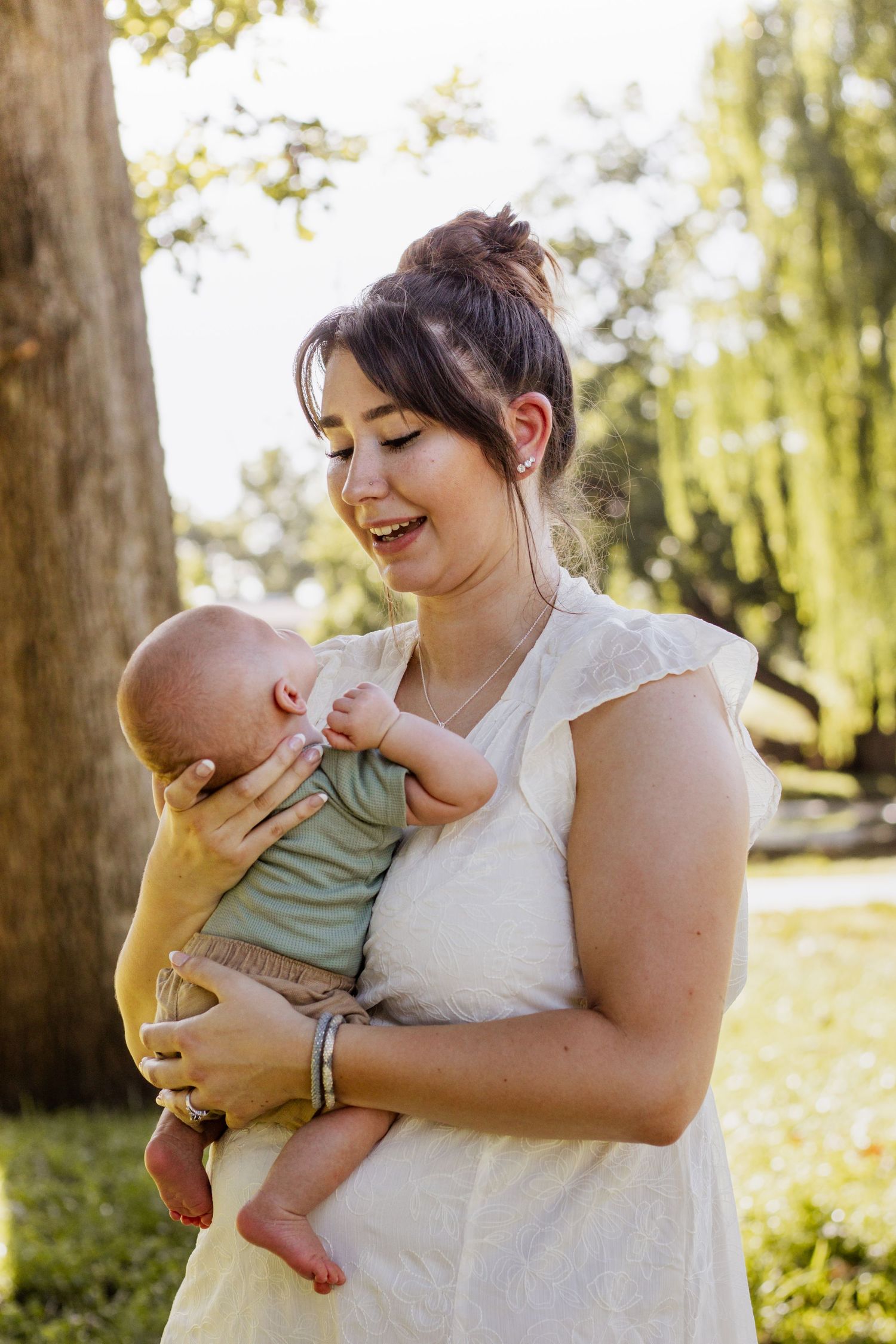 Smiling young mother in white dress holding her newborn baby outdoors in a sunny green park setting.