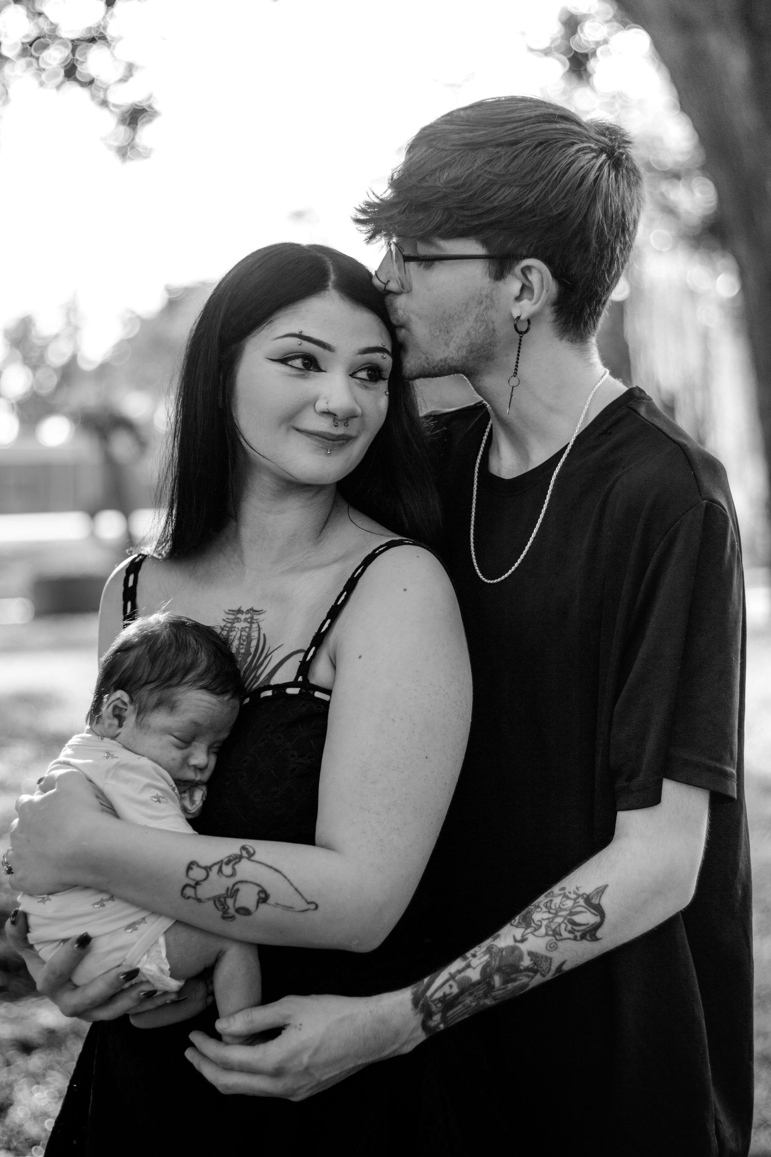 Black and white photo of tattooed couple with newborn baby outdoors, man kissing womans cheek.