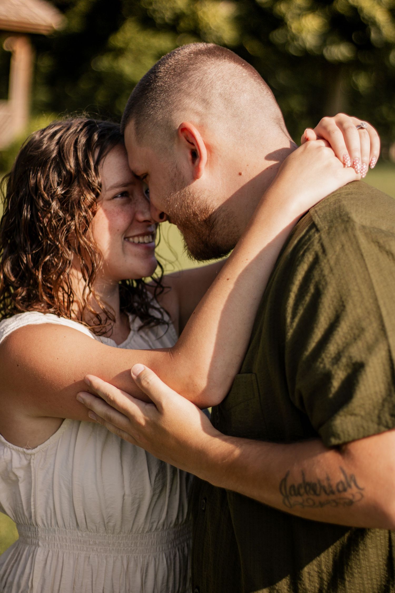 Couple sharing a tender forehead touch outdoors, smiling woman in white dress embracing man in green shirt.