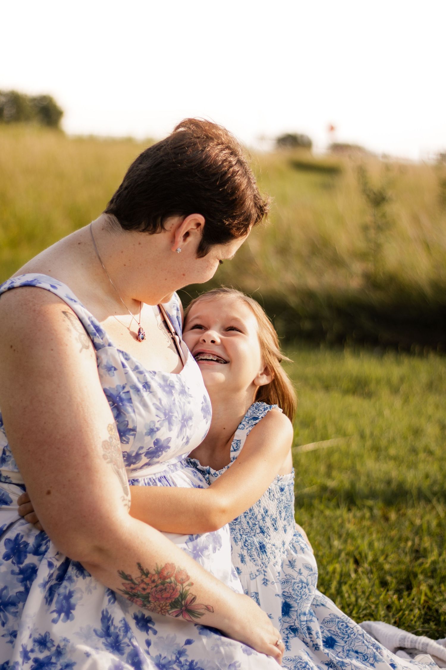 A mother and daughter in matching blue floral dresses share a joyful moment sitting together in a sunlit green field.
