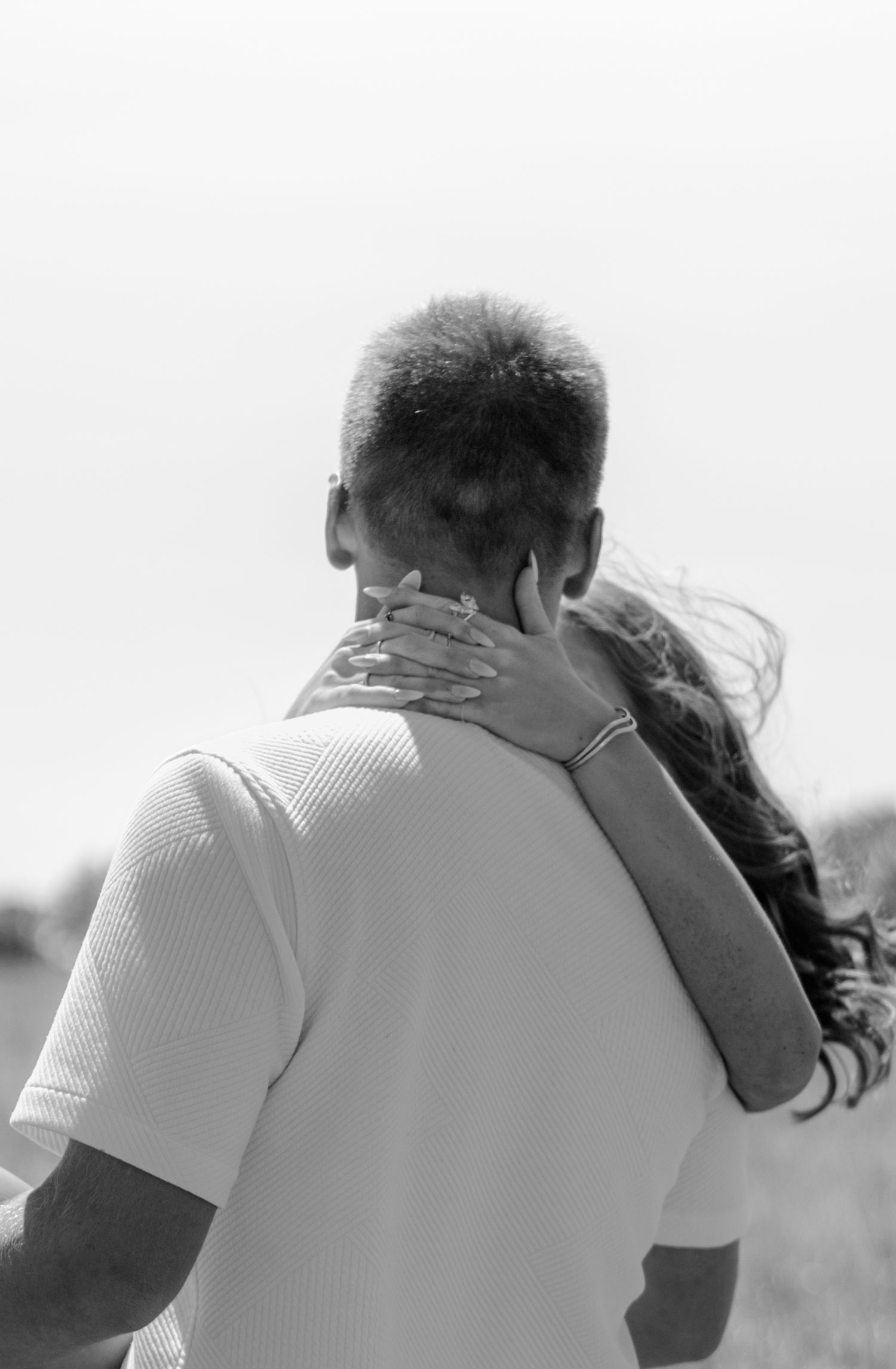 Black and white photo of a couple embracing outdoors, woman's hand resting on man's neck, hair blowing in wind.