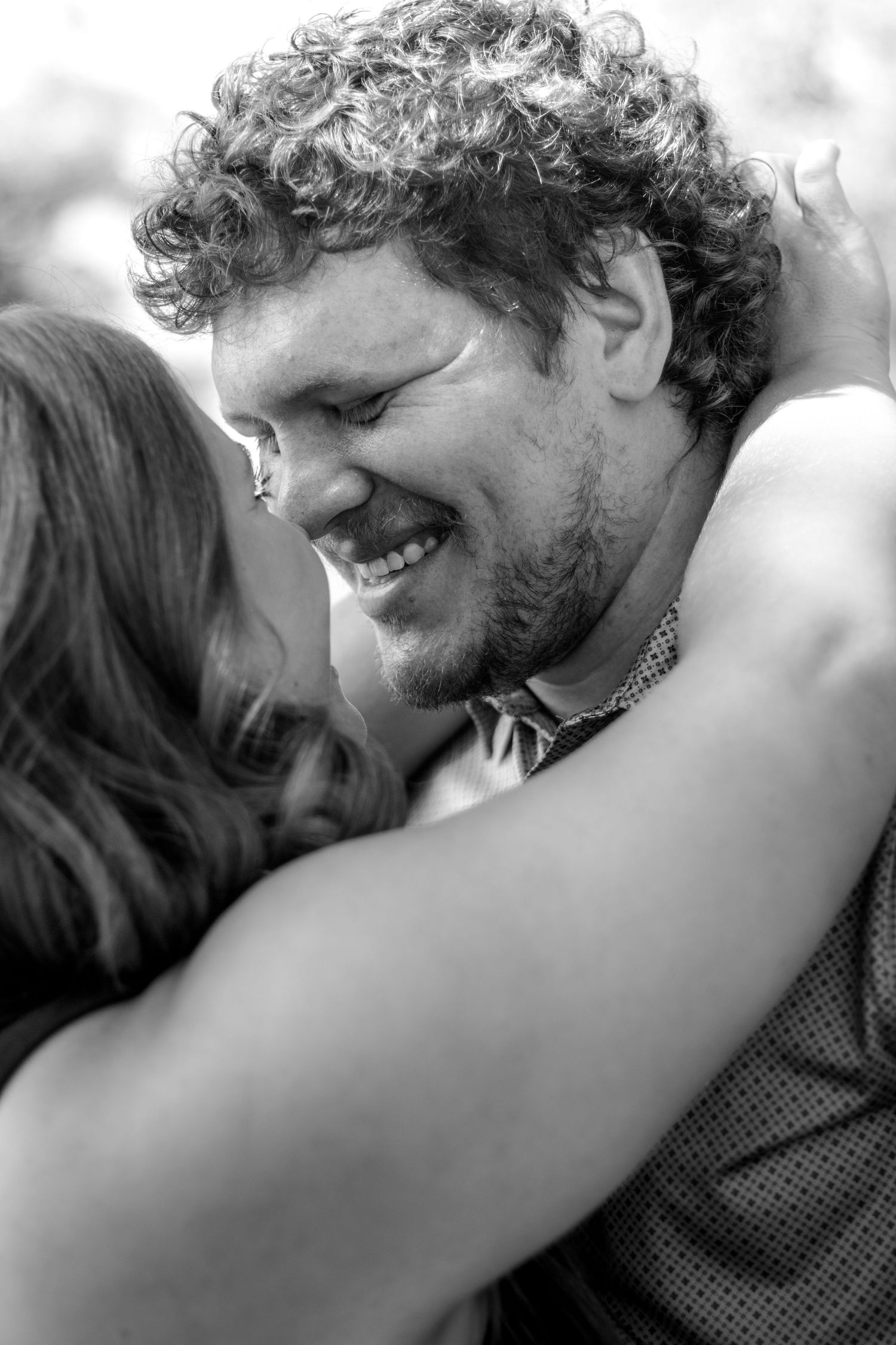Black and white photo of a smiling curly-haired man being embraced by a woman, sharing a joyful moment.