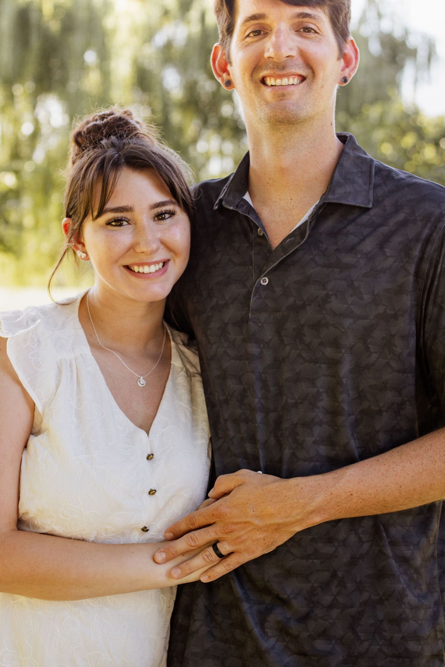Smiling couple posing outdoors, woman in white dress and man in dark polo shirt, standing together in a sunny park.