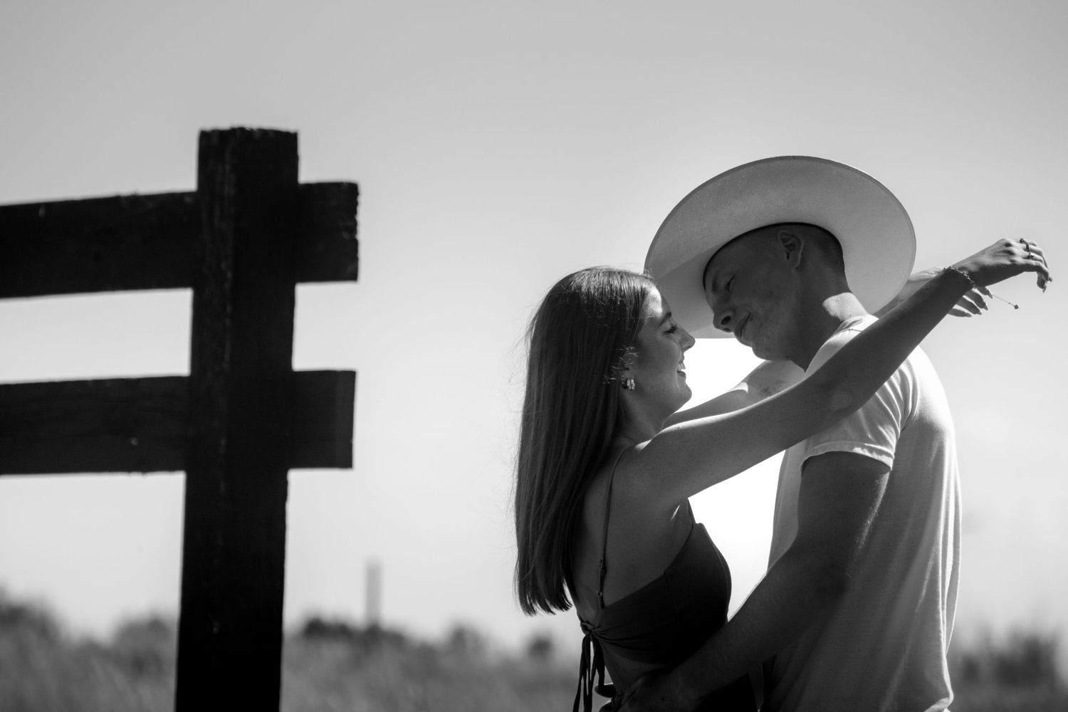 Black and white photo of a couple sharing a romantic moment near a wooden cross in an outdoor setting.