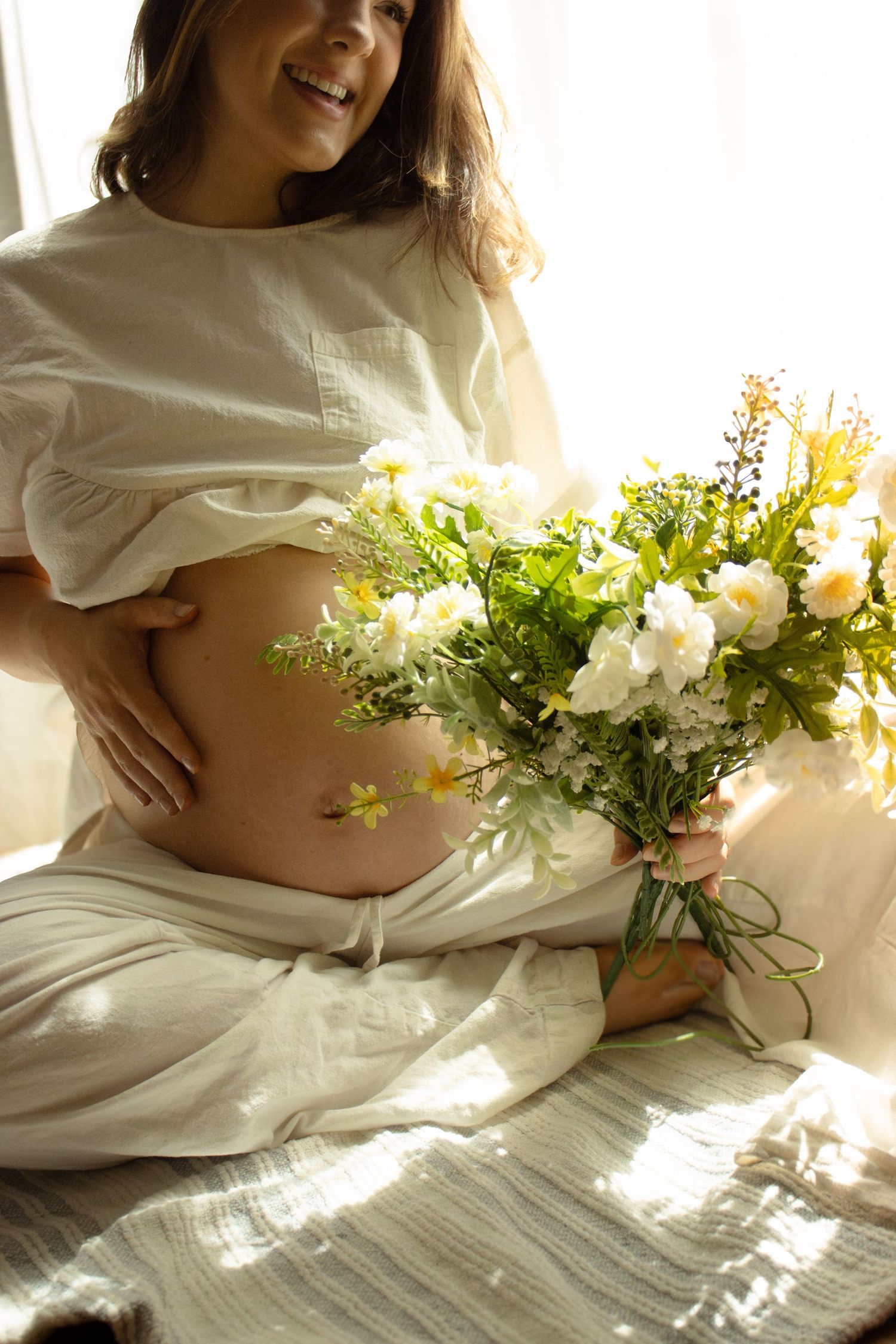 Happy pregnant woman in white top holding colorful wildflower bouquet, cradling her bare belly in soft natural light.