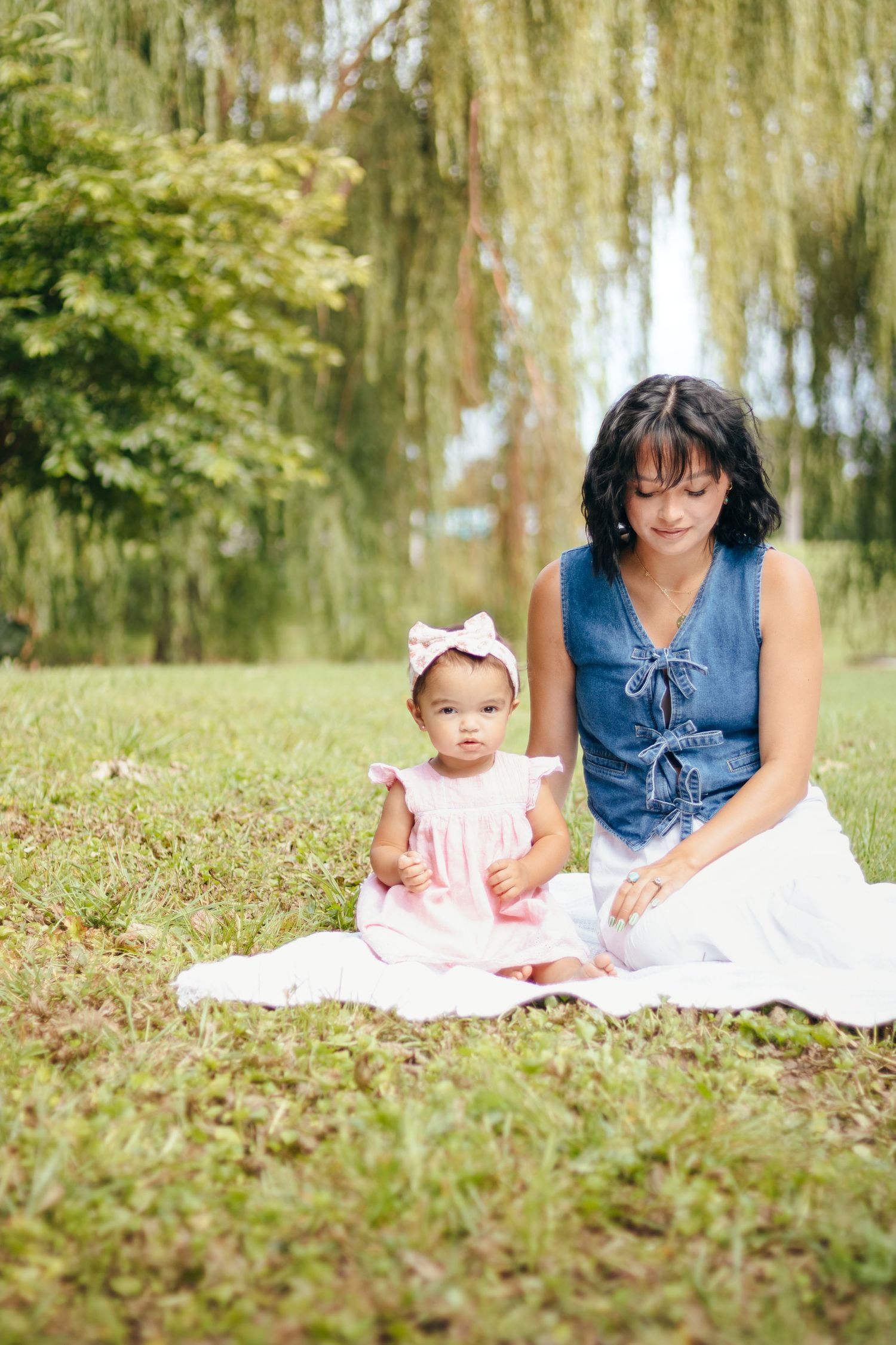Mother and baby girl sitting on blanket in green park, baby wearing pink dress with bow headband.