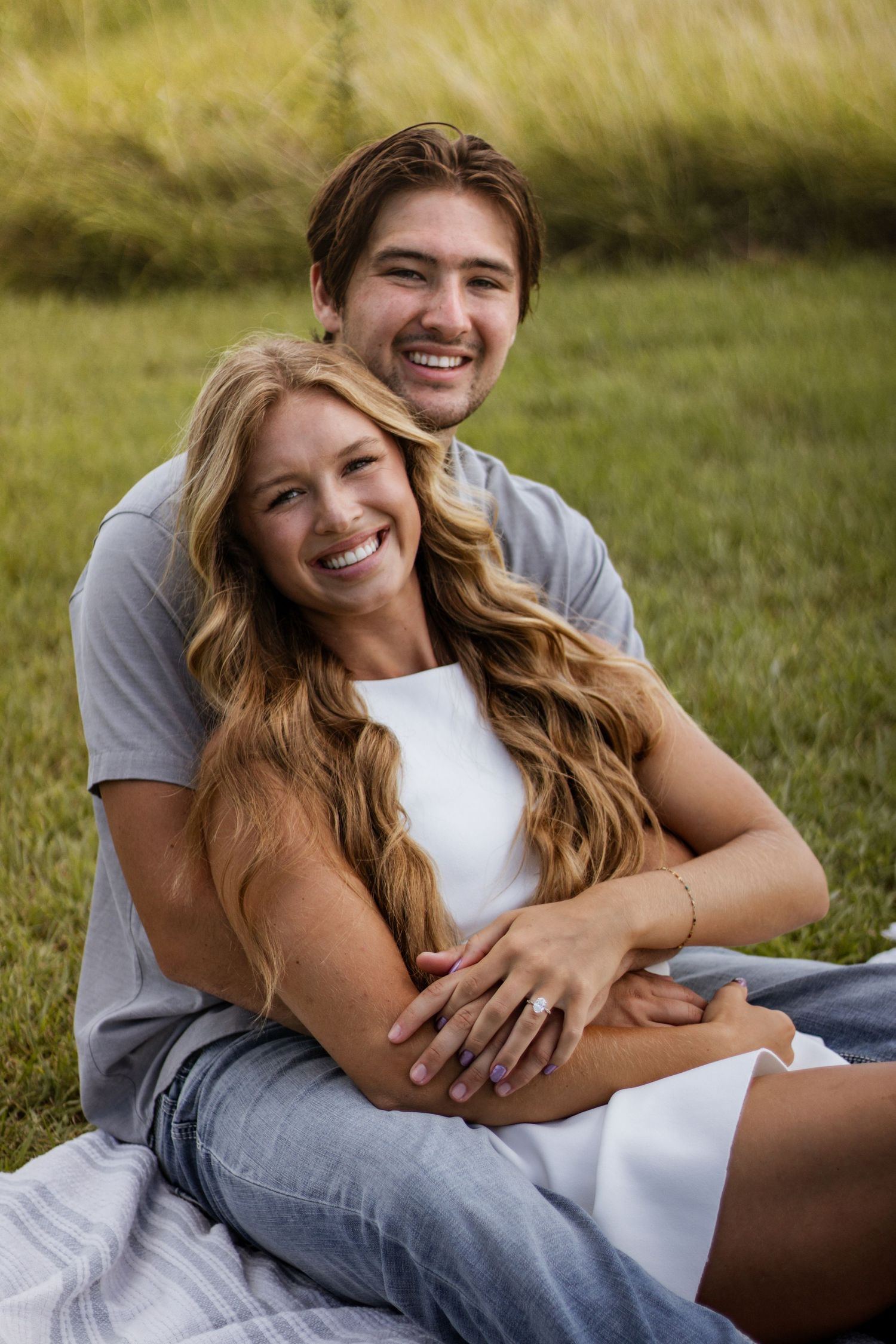 Smiling couple sitting together in green grass, man embracing woman from behind in outdoor photo.