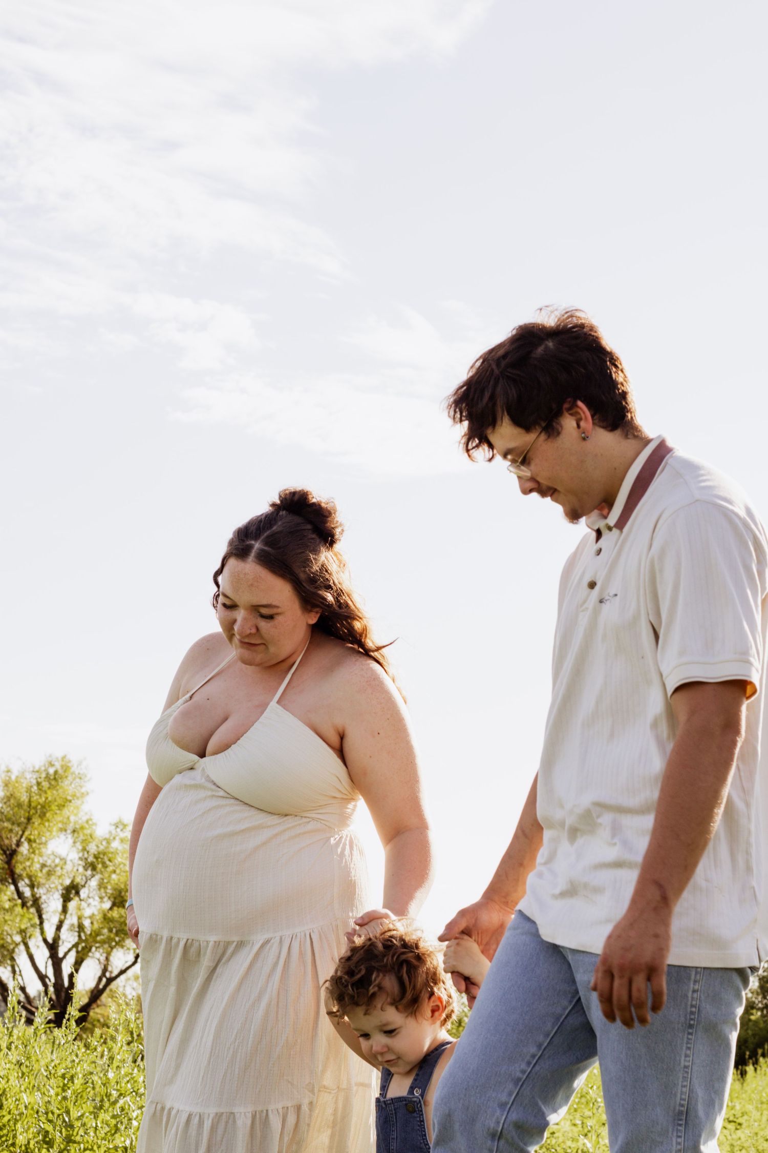 Pregnant woman and man hold toddler's hands outdoors in sunny green field, family maternity photo session.