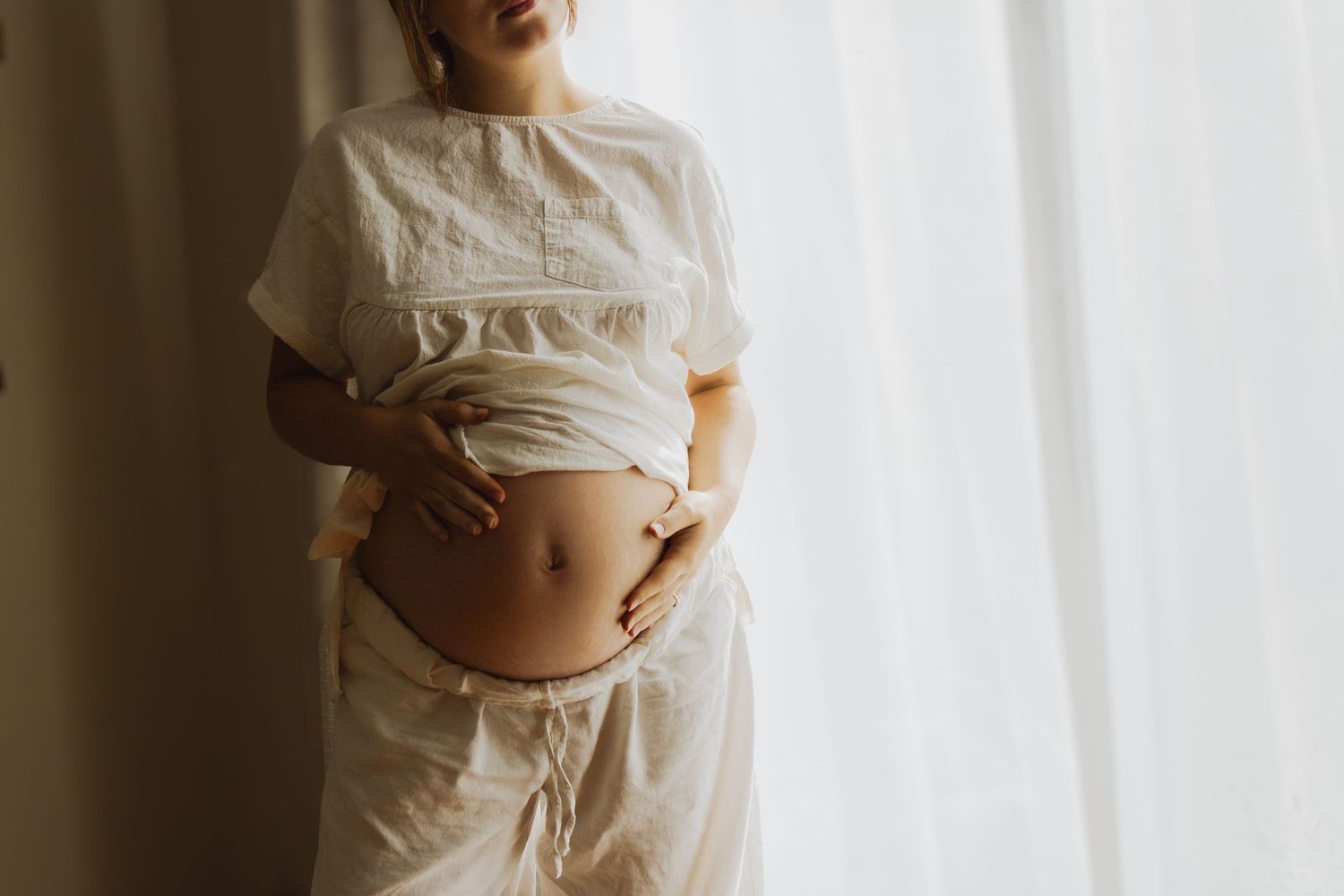 Pregnant woman in white linen top holding her bare belly near a bright window.