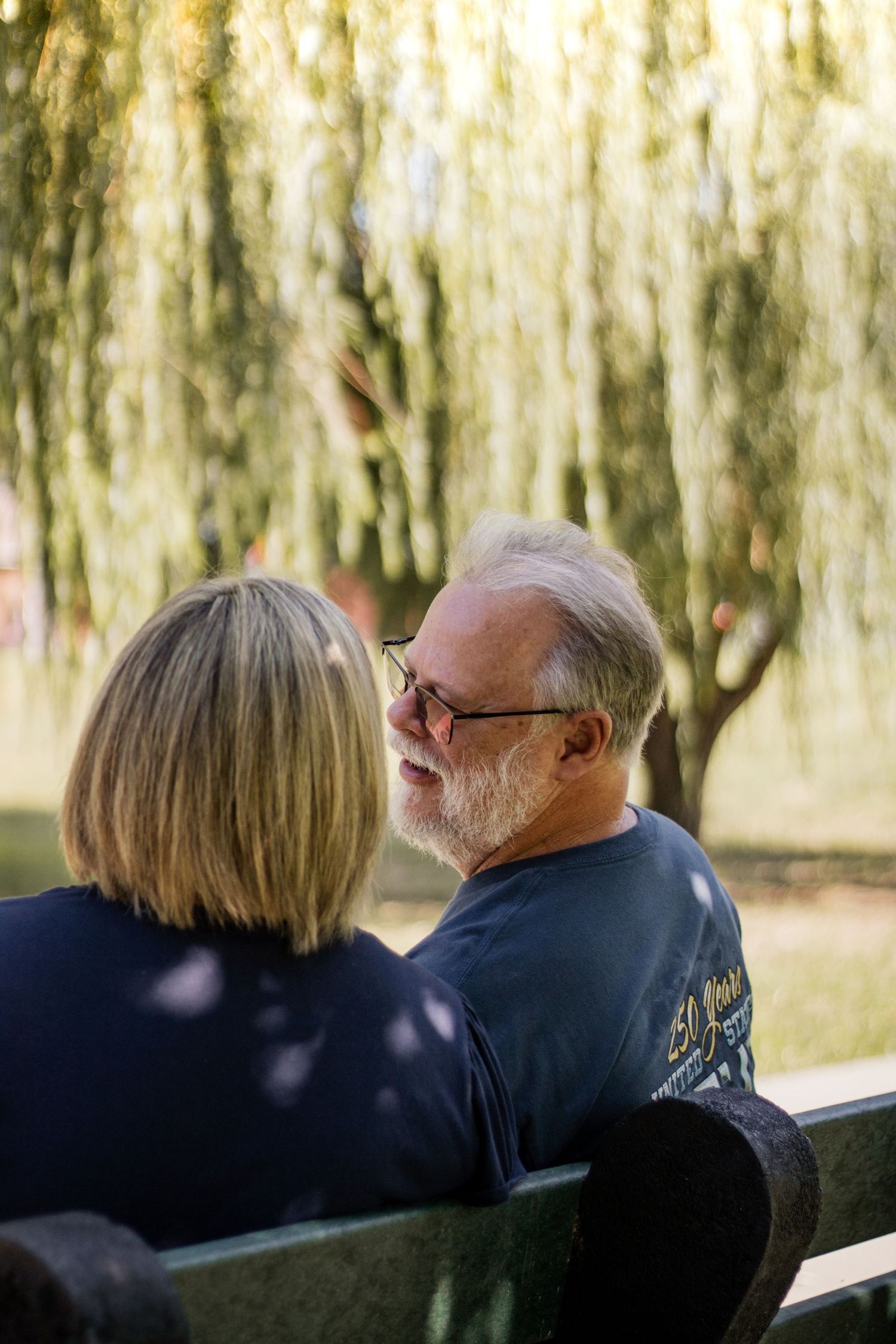Elderly couple sitting on a park bench conversing, with weeping willow trees in the background on a sunny day.