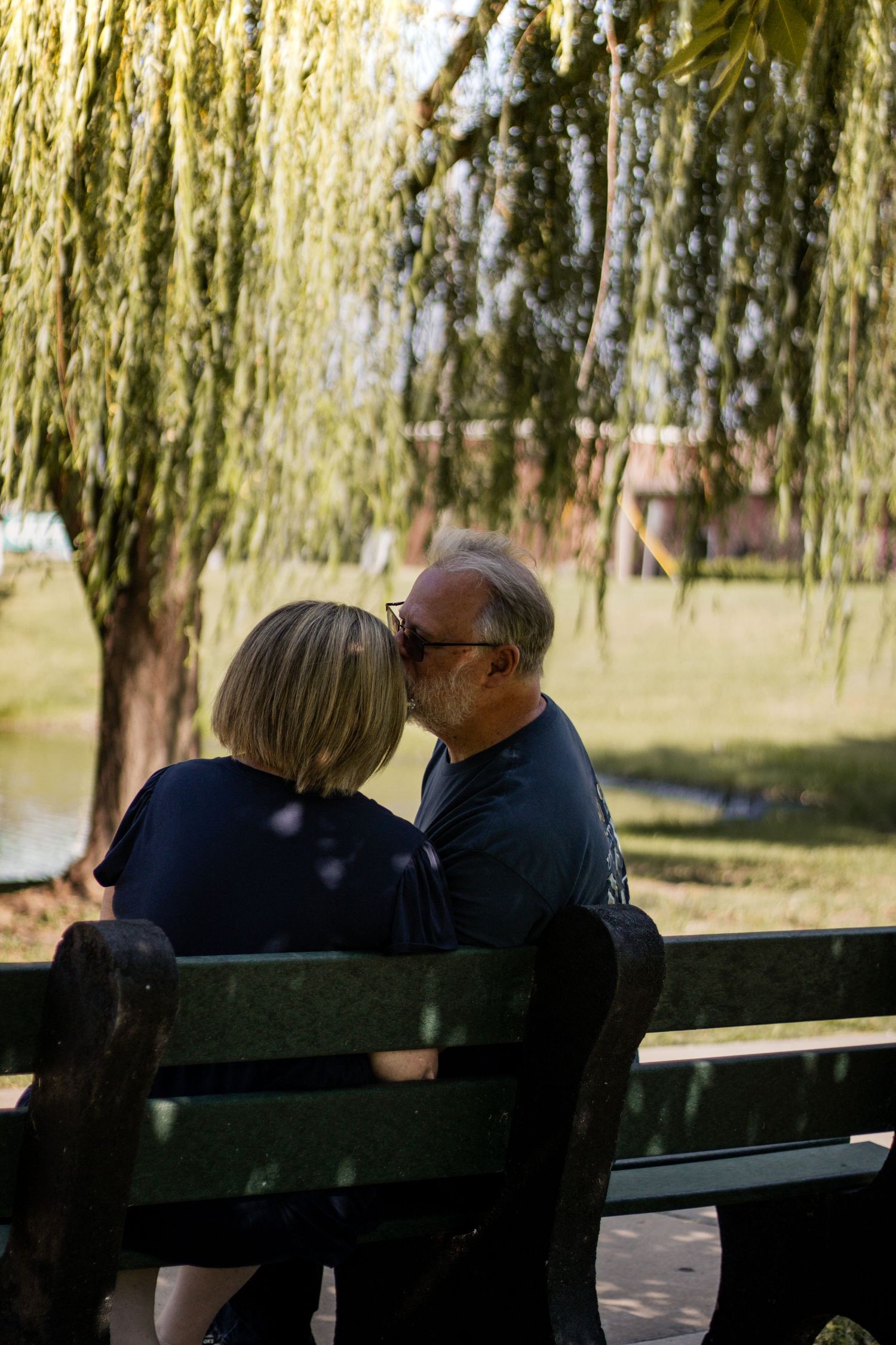 Couple sharing a romantic moment on a park bench under a weeping willow tree by a pond.