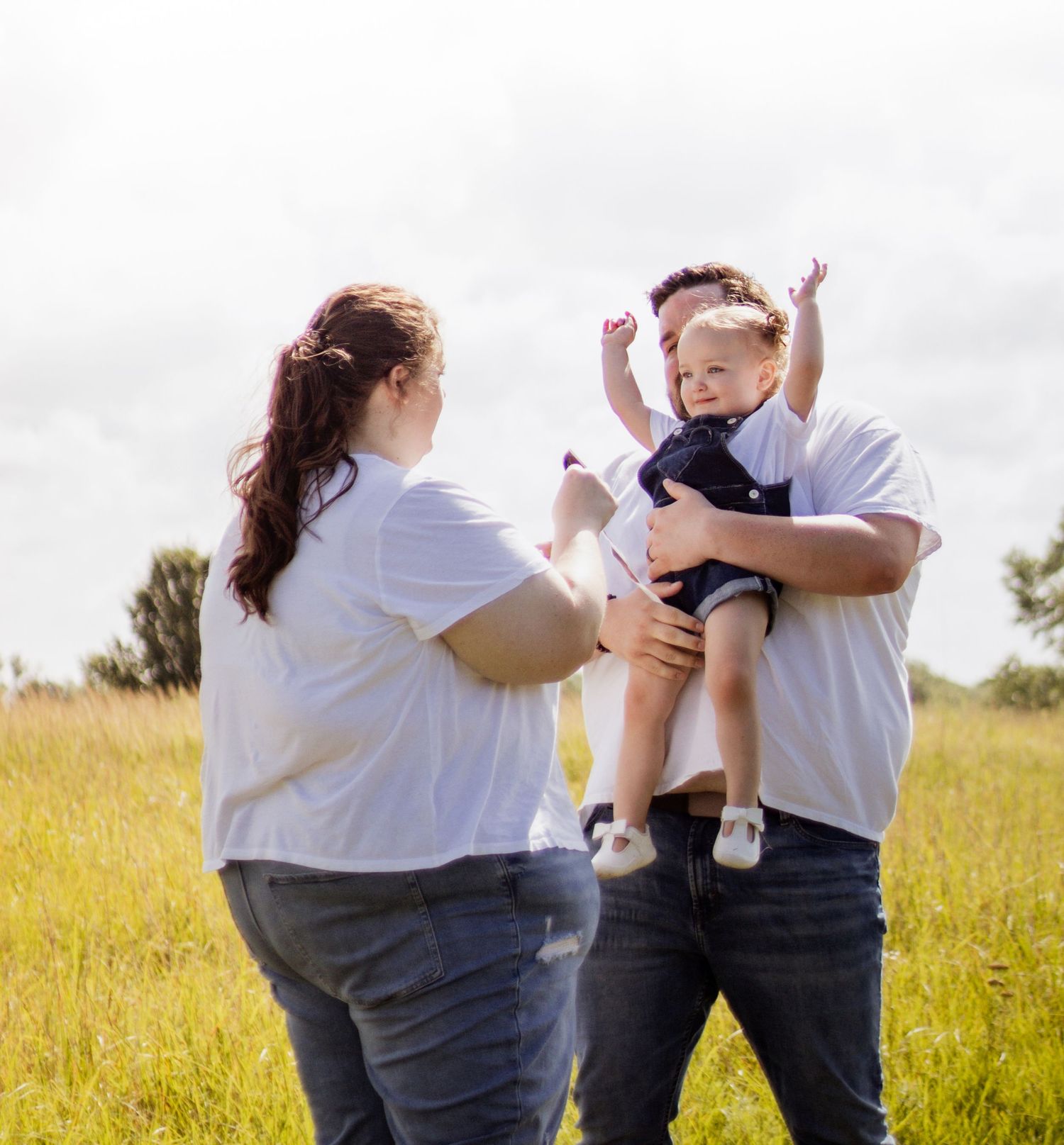A joyful family in white shirts lifts a happy baby outdoors in a sunny green field.