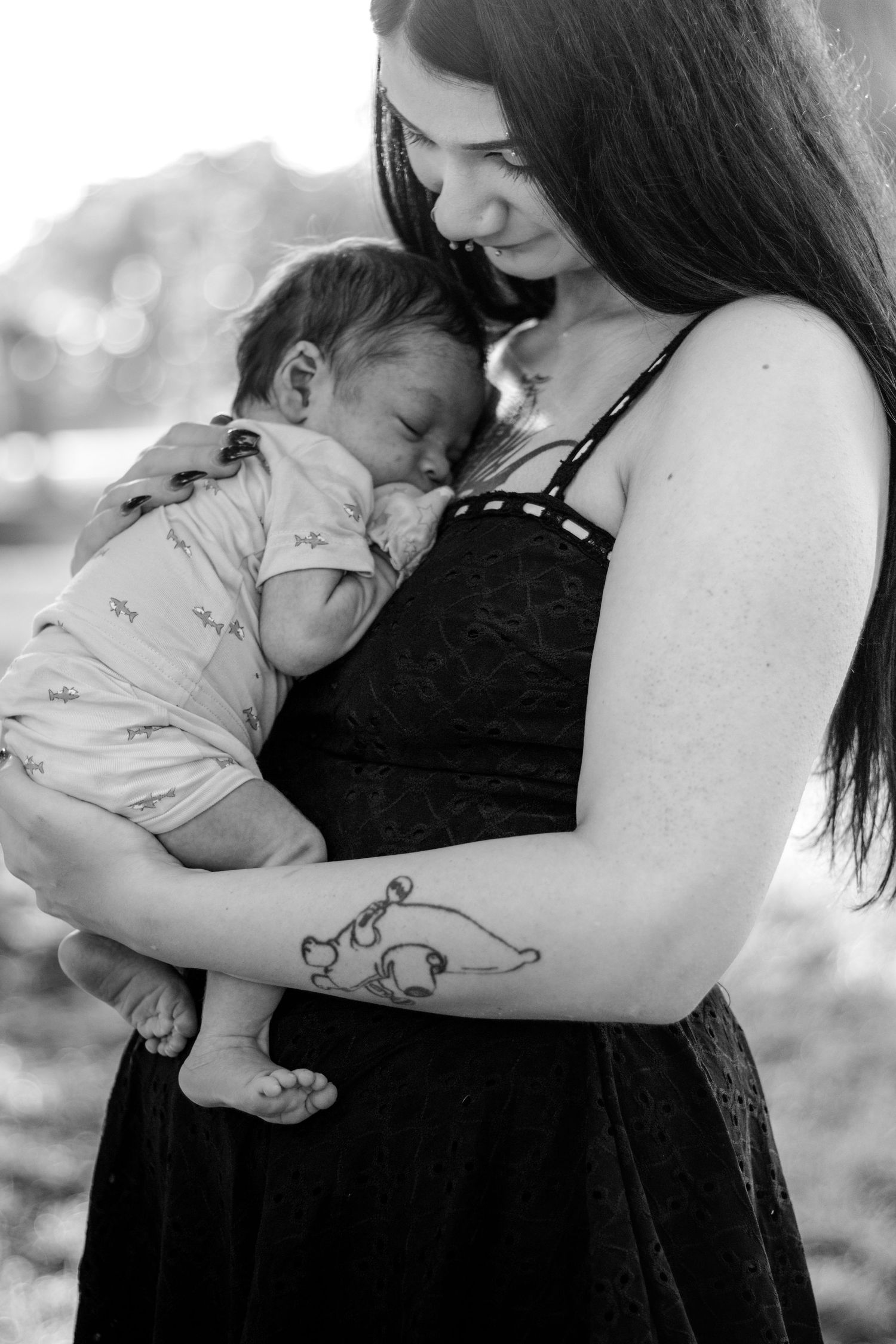 Black and white photo of a mother with tattoos cradling her sleeping newborn baby outdoors.